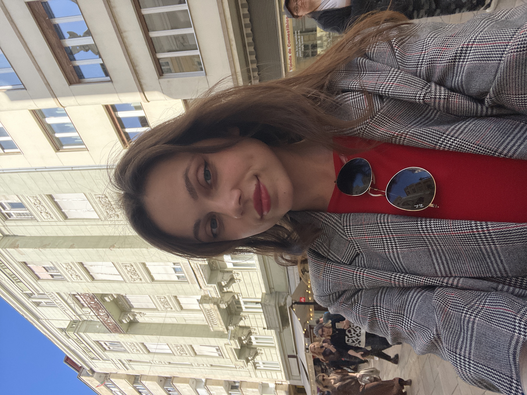 A woman is taking a selfie in Budapest, Hungary. She is wearing a grey plaid blazer over a red top, and sunglasses are resting on her chest. A building with multiple windows is behind her, and a sign reading "Tienda de Comestibles" can be seen. A crowd of people is in the background. The sky is blue.