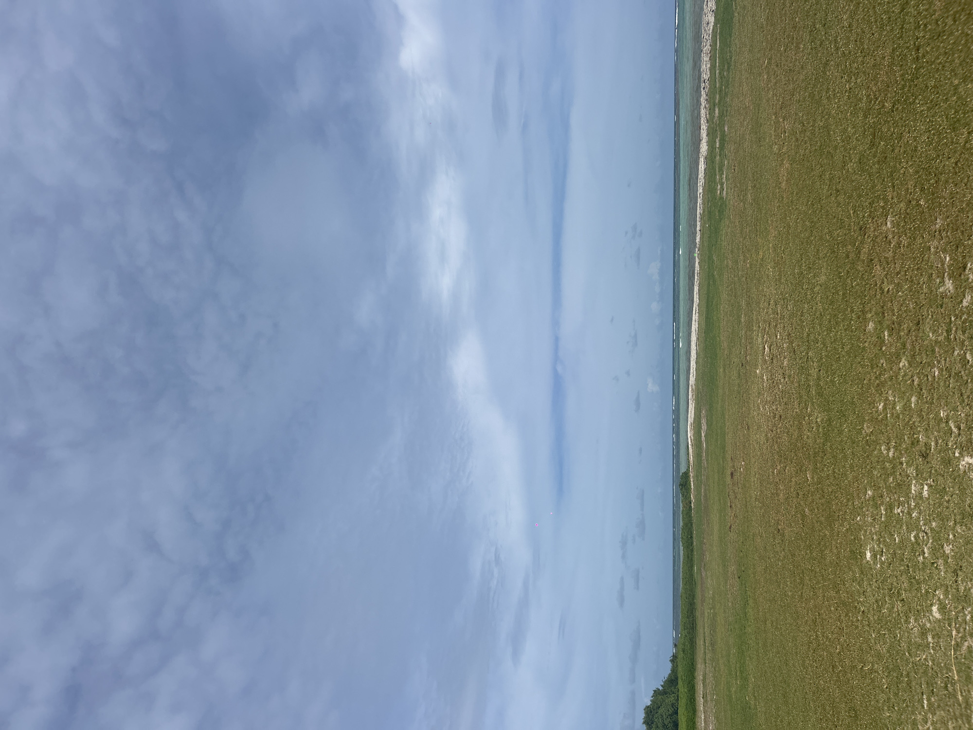 The image depicts a coastal scene near Sainte-Anne, France. A wide expanse of sky, filled with layered clouds, dominates the left side. The ocean, with a narrow strip of white sand beach, runs along the center. To the right, a field of grassy vegetation stretches towards the edge of the frame. A line of low-lying shrubs and trees separates the field from the beach. No people, specific objects, or activities are visible. The scene is characterized by natural elements: sky, ocean, beach, vegetation.