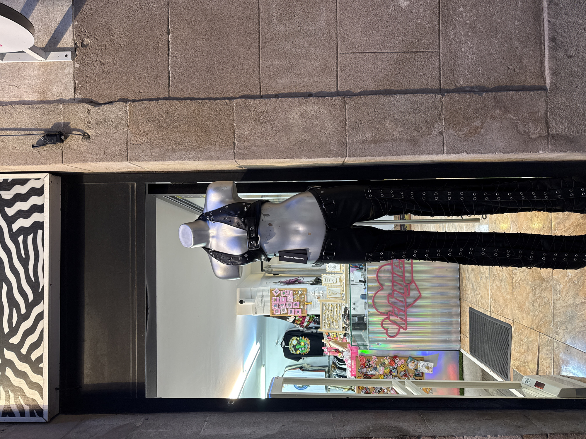 A storefront in Barcelona, Spain, is viewed from the exterior through its glass window. A mannequin is dressed in black leather clothing with metal accents; it is positioned inside the store, facing outwards. The store's interior features displays of clothing, accessories, and various merchandise. Outside, the building's facade consists of large, rectangular stone blocks. The scene is illuminated by the store's interior lighting and ambient city light.