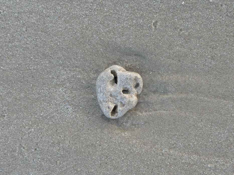 A light gray, irregularly shaped stone, approximately 8-10 cm across its widest point, is partially embedded in fine-grained, damp, light beige sand. The stone features three naturally eroded cavities on its visible surface: two superior, horizontally elongated depressions resembling eye sockets, and one inferior, roughly circular opening consistent with a nasal aperture. The stone's texture is uneven and porous. Faint, dark, vertical striations are present in the sand directly below the stone, suggesting subtle water movement or prior disturbance. The scene is observed from an overhead perspective, depicting a static beach environment in Tel-Aviv, Israel. No other subjects or objects are visible within the frame.