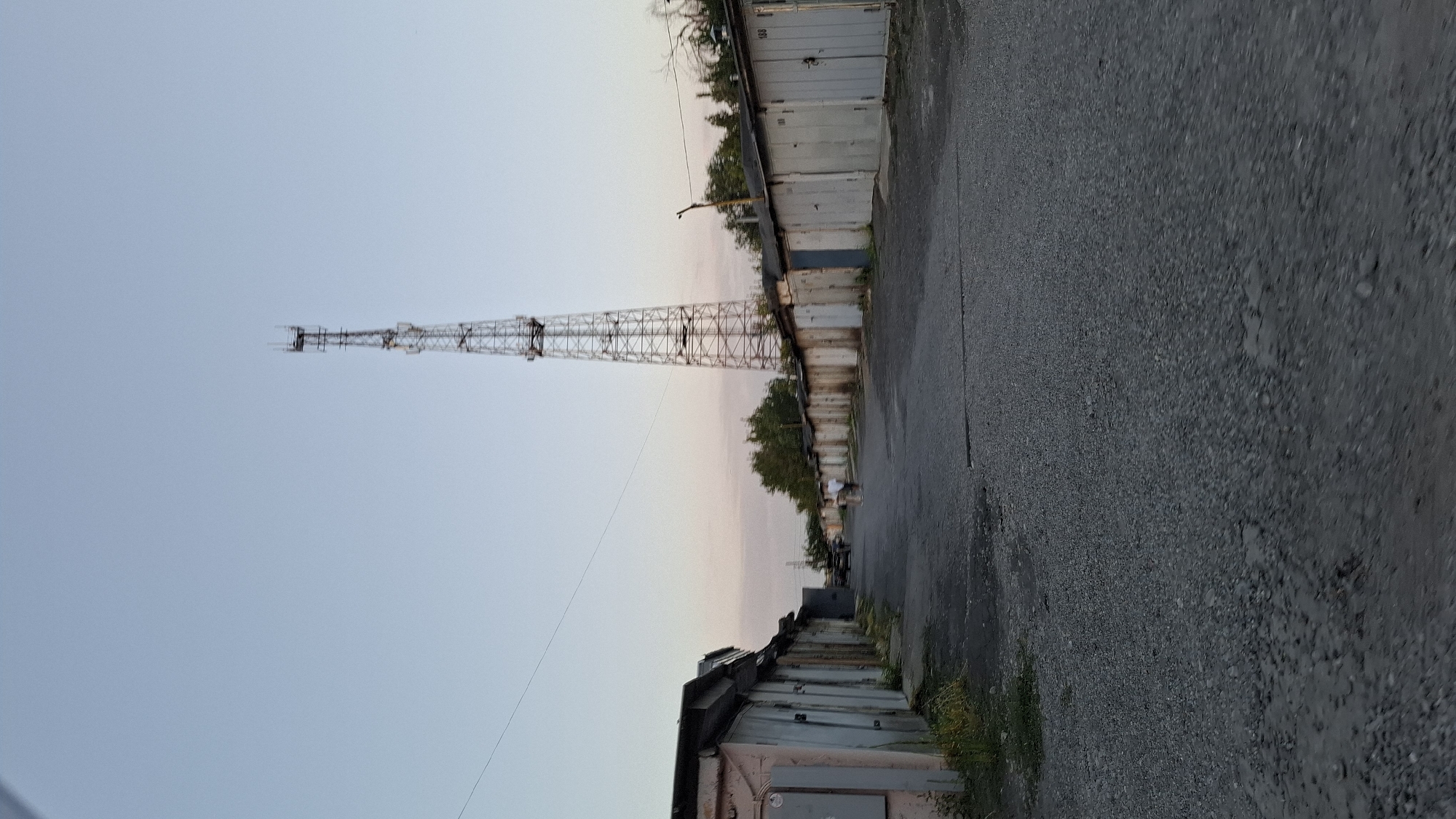 An exterior, vertical, full shot captures a view down a gravel road lined with garages on both sides, leading towards a distant cell tower. The sky is a clear, pale blue with a faint pinkish hue near the horizon, indicating either sunrise or sunset. In the middle distance, two individuals, appearing to be a man and a woman, are walking away from the camera down the road. To the left, a row of garages is visible, some with their doors closed and others partially open, with a few patches of greenery growing along their bases. To the right, another row of garages extends into the distance, with some numbered doors visible. The cell tower is a tall, lattice structure with antennas at its apex. Trees are visible beyond the garages on the right side. The scene is likely set in a residential or industrial area on the outskirts of Shymkent, Kazakhstan, characterized by utilitarian structures and open spaces.