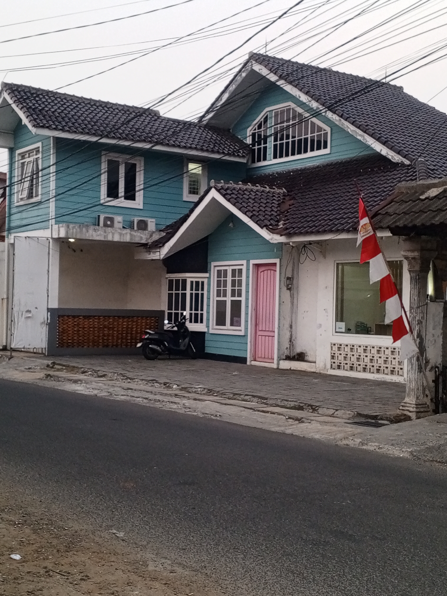 The image captures a two-story building with a teal facade on the upper level and white on the lower level, situated in Depok, Indonesia. The building features a dark, tiled roof. The front of the building has several windows, including a large arched window on the upper floor with white framing, and a pink door with matching window frames on the lower floor. A black motorcycle is parked on the paved area in front of the building. To the right, a white section of the building is visible with a window and decorative ventilation blocks below it. A red and white flag is prominently displayed on a pole to the right of the building. The scene is set against an overcast sky, with power lines visible crisscrossing overhead. The foreground shows a paved road and sidewalk.