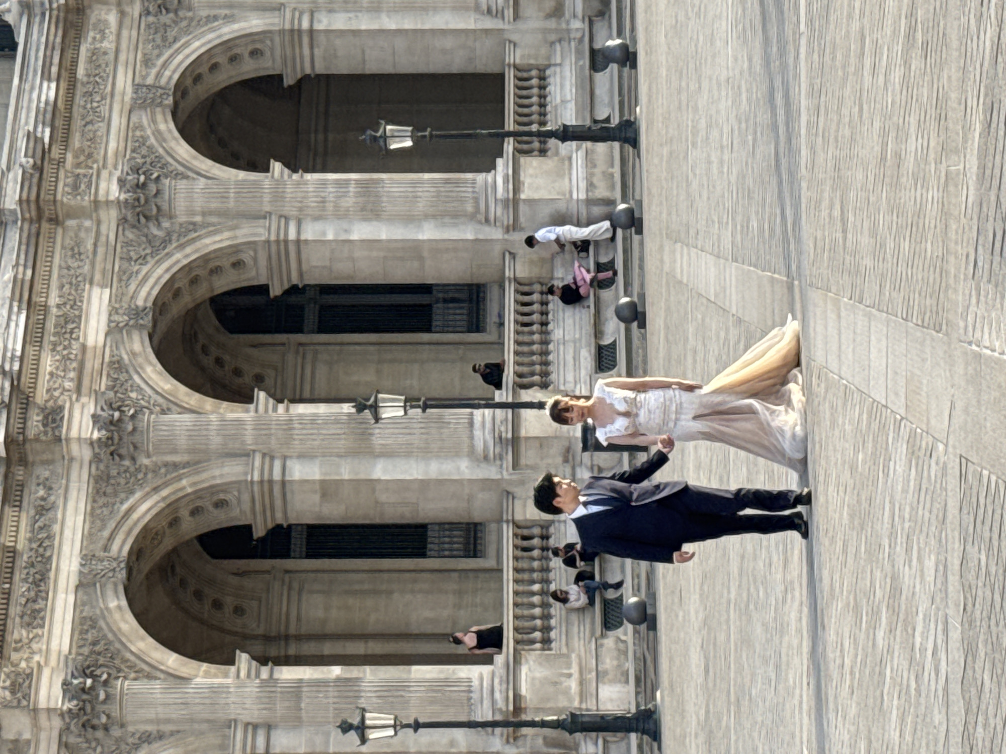 A recently married couple, the bride in an ivory gown with a champagne sash and the groom in a dark suit, poses for a photograph with the Palais Garnier, a 19th-century opera house in Paris, France, visible in the background. Several other people are visible in the background, along with multiple historical street lamps.