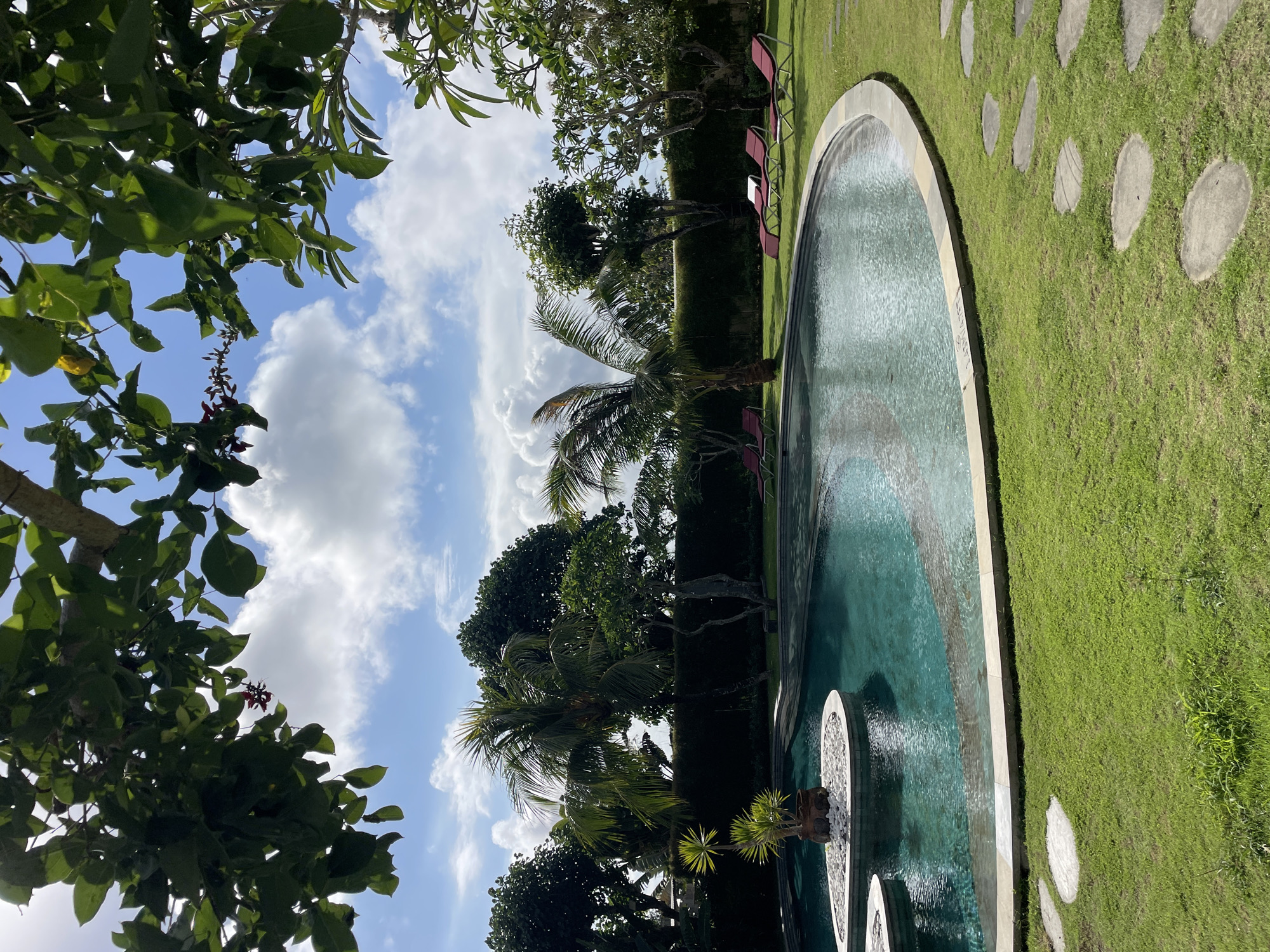 The photograph depicts an outdoor swimming pool surrounded by lush greenery and a partially visible sky. The pool is turquoise in color and appears to be either round or oval. Adjacent to the pool is a well-maintained grassy area punctuated by large, circular stepping stones.
Several red chairs are positioned near the edge of the pool, suggesting an area for relaxation. The foreground and background of the image are dominated by various trees and foliage. The sky is visible with white clouds and blue patches.
The location appears to be a private garden or a resort in a tropical or subtropical climate. The city and region are unknown.