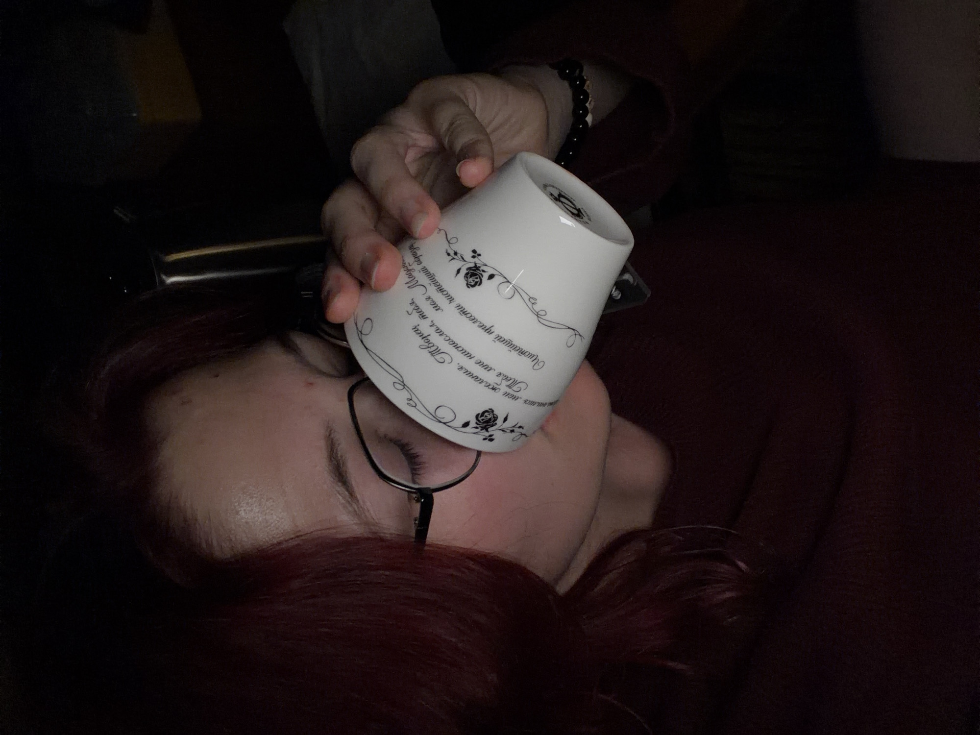 The image shows a close-up of a person with reddish-brown hair and glasses drinking from a white mug. The mug has decorative text and floral patterns on it. The person is wearing a dark red sweater. The background is dark and out of focus, suggesting an indoor setting at night or in low light. There is no information in the image to suggest a location in Chelyabinsk, Russia.