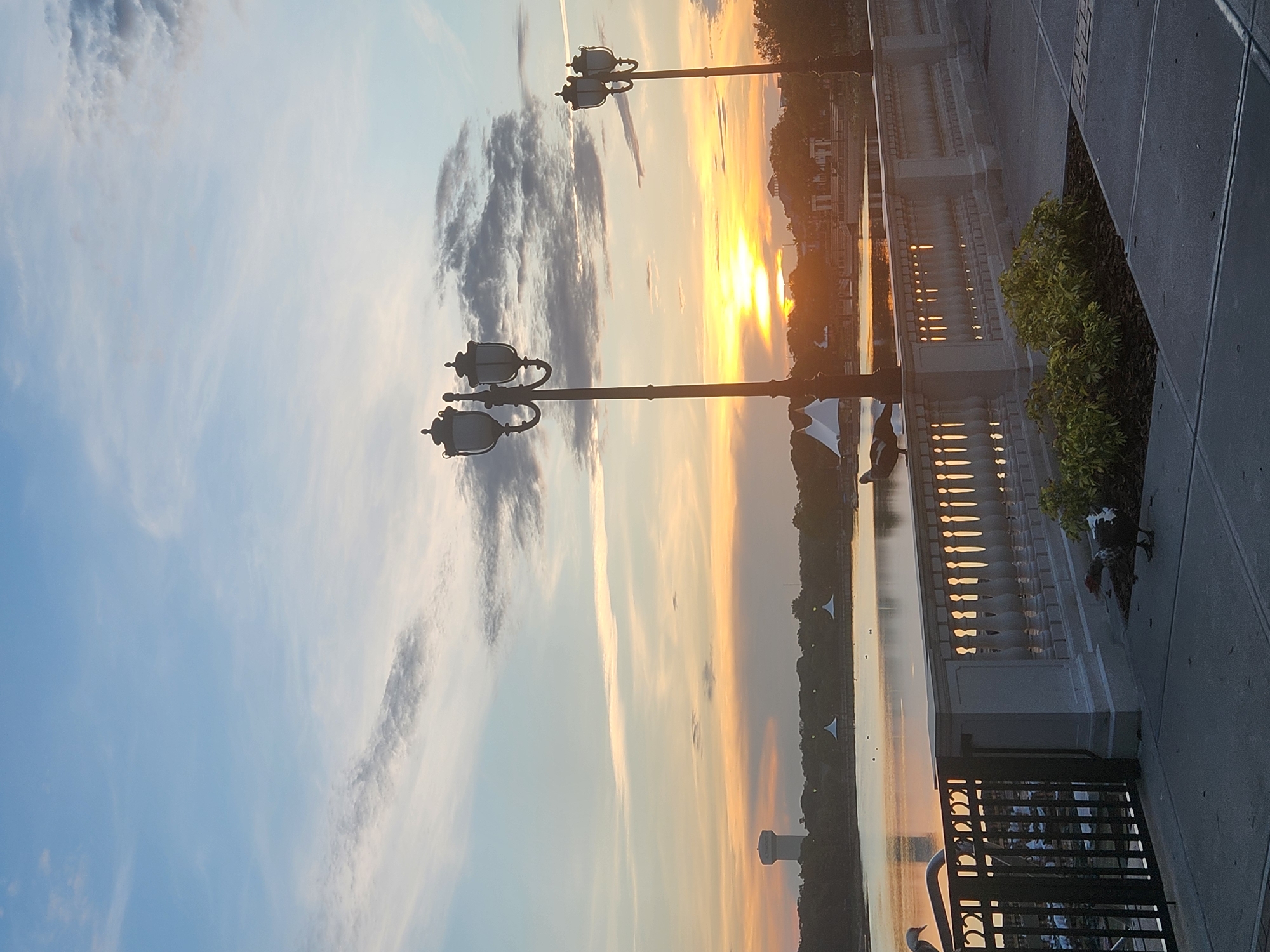 This is a photograph taken at Crane's Roost Park in Altamonte Springs, Florida, USA, during a sunset. The scene captures a tranquil waterfront promenade. In the foreground, there are two ornate lampposts on the right and a black metal fence on the left. A white stone balustrade runs along the edge of the promenade, overlooking the water. Two ducks are visible, one perched on the balustrade and another on the ground near some greenery. The water of Crane's Roost Lake reflects the warm colors of the sunset. In the distance, a water tower stands on the left, and trees and some white tent-like structures are visible along the shoreline. The sky is a blend of soft blue with streaks of orange and yellow from the setting sun, punctuated by scattered clouds. The overall atmosphere is peaceful and serene, characteristic of a park at dusk.