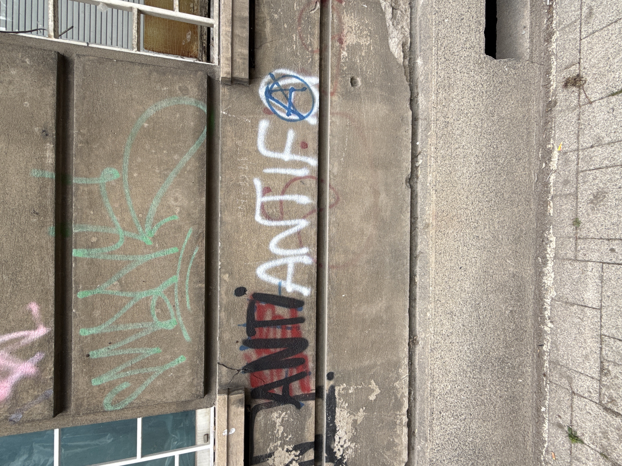 The image shows graffiti on the exterior wall of a building in Sofia, Bulgaria. The graffiti consists of words and symbols spray-painted in various colors.
In the upper section of the image, green graffiti is visible on a concrete surface. Below this, a prominent phrase appears in black and white spray paint, with some red and blue accents. The words "ANTIFA" are clearly legible in large, blocky letters. To the right of "ANTIFA," a blue circle with an "A" inside, a symbol associated with anarchism, is also spray-painted. Faint red lines and circles are visible beneath the "ANTIFA" text.
The building's facade is made of rough concrete. A section of a window with white framing and blue plastic sheeting is visible on the left side of the image. On the right, part of another window with a metal grate and amber-colored panes can be seen.
The lower part of the image shows the ground level of the building, which is also concrete, and a sidewalk made of stone pavers. A dark, rectangular opening, possibly a ventilation grate or drain, is present in the lower right portion of the wall. The overall scene suggests an urban environment where graffiti is present on public or semi-public surfaces.