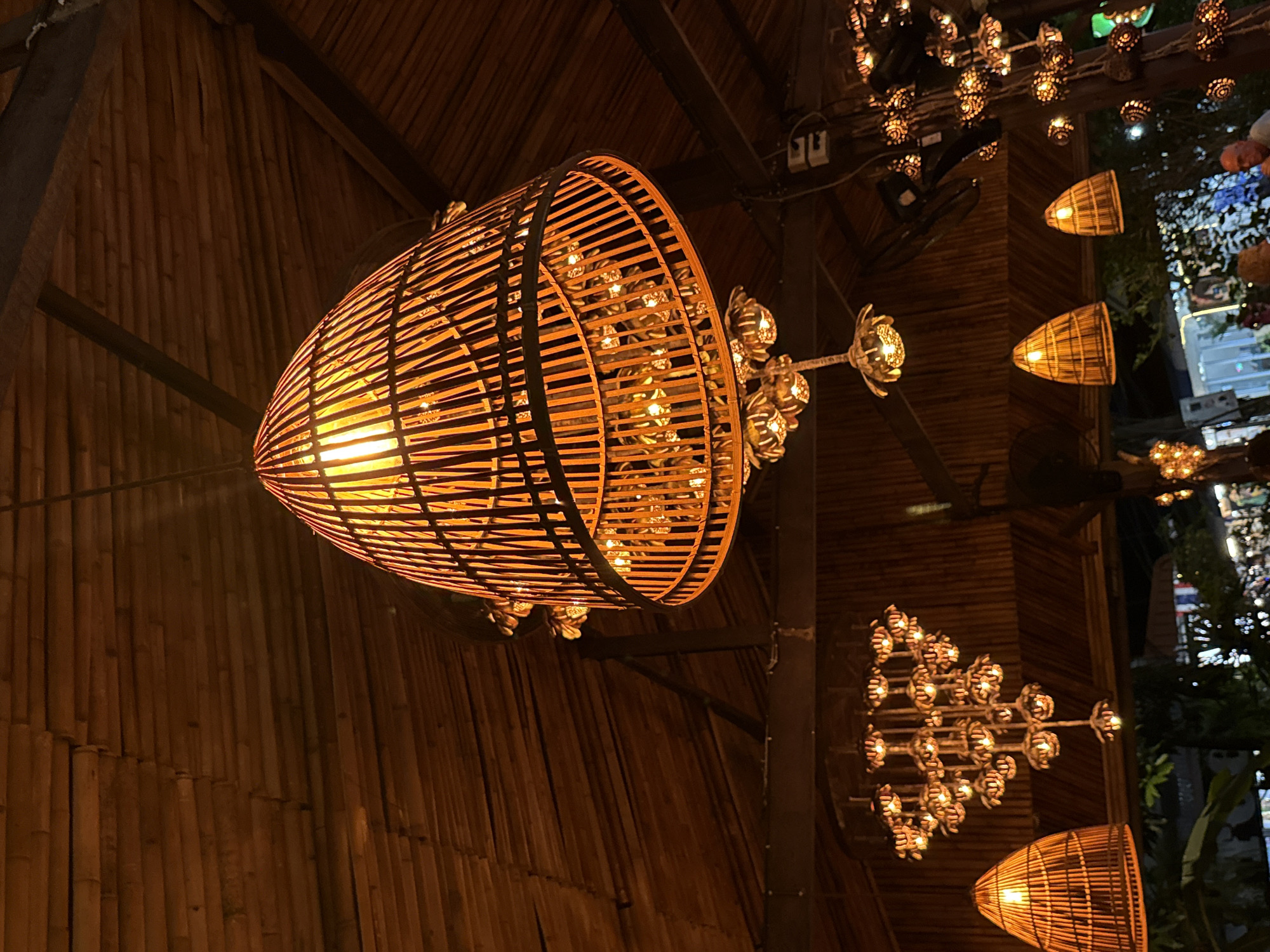 This image captures a warmly illuminated interior or covered outdoor setting, characterized by natural materials and ambient lighting, likely at night. The central focus is a large, elongated lantern, intricately crafted from light-colored bamboo or wooden strips, forming a cage-like structure. It emits a bright, warm orange glow from within, serving as a primary light source.
The setting features a ceiling and walls paneled with vertical bamboo stalks, giving the space a rustic and natural aesthetic. Dark wooden beams and structural elements are visible overhead, from which the lanterns are suspended. In the background and to the right, several other smaller lanterns are visible. Some are individual pendant lights, similar in style to the main lantern, while others are clustered into elaborate, multi-faceted fixtures resembling chandeliers, all casting a harmonious warm light.
Beyond the immediate structural elements, the background is softly blurred and dark, revealing glimpses of outdoor foliage and scattered artificial lights, suggesting an adjacent outdoor area or a view into the distance. The overall impression is one of a cozy, inviting, and exotic atmosphere, predominantly lit by the golden hues of the lanterns. No people or visible text are present. The decor and materials strongly suggest a tropical or resort-like environment, consistent with the location of Baan Lamai, Thailand.