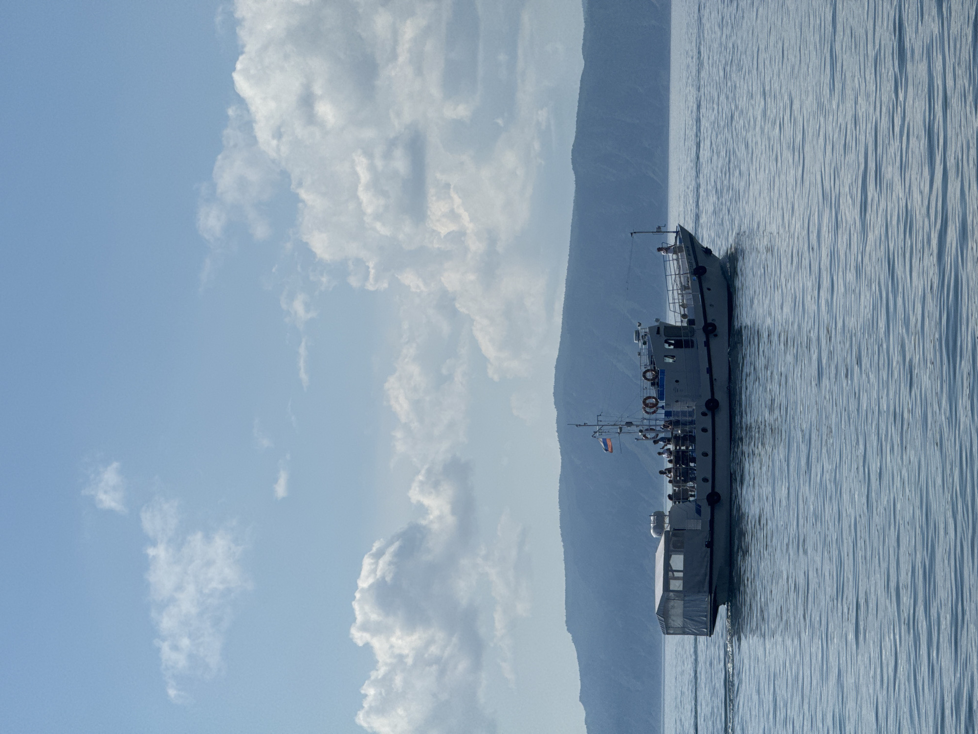 In Listvyanka, Russia, a small, white boat with a covered seating area moves across a calm body of water. The boat is populated with approximately six to eight individuals visible on its deck, some standing and others seated. A Russian flag flies from a mast at the rear of the boat. In the background, a large, blue-hued mountain range stretches across the horizon. The sky above is a clear blue, interspersed with fluffy white clouds. The water is a muted blue, with gentle ripples indicating movement. The scene suggests a clear, sunny day, likely during a tour or excursion on the water.