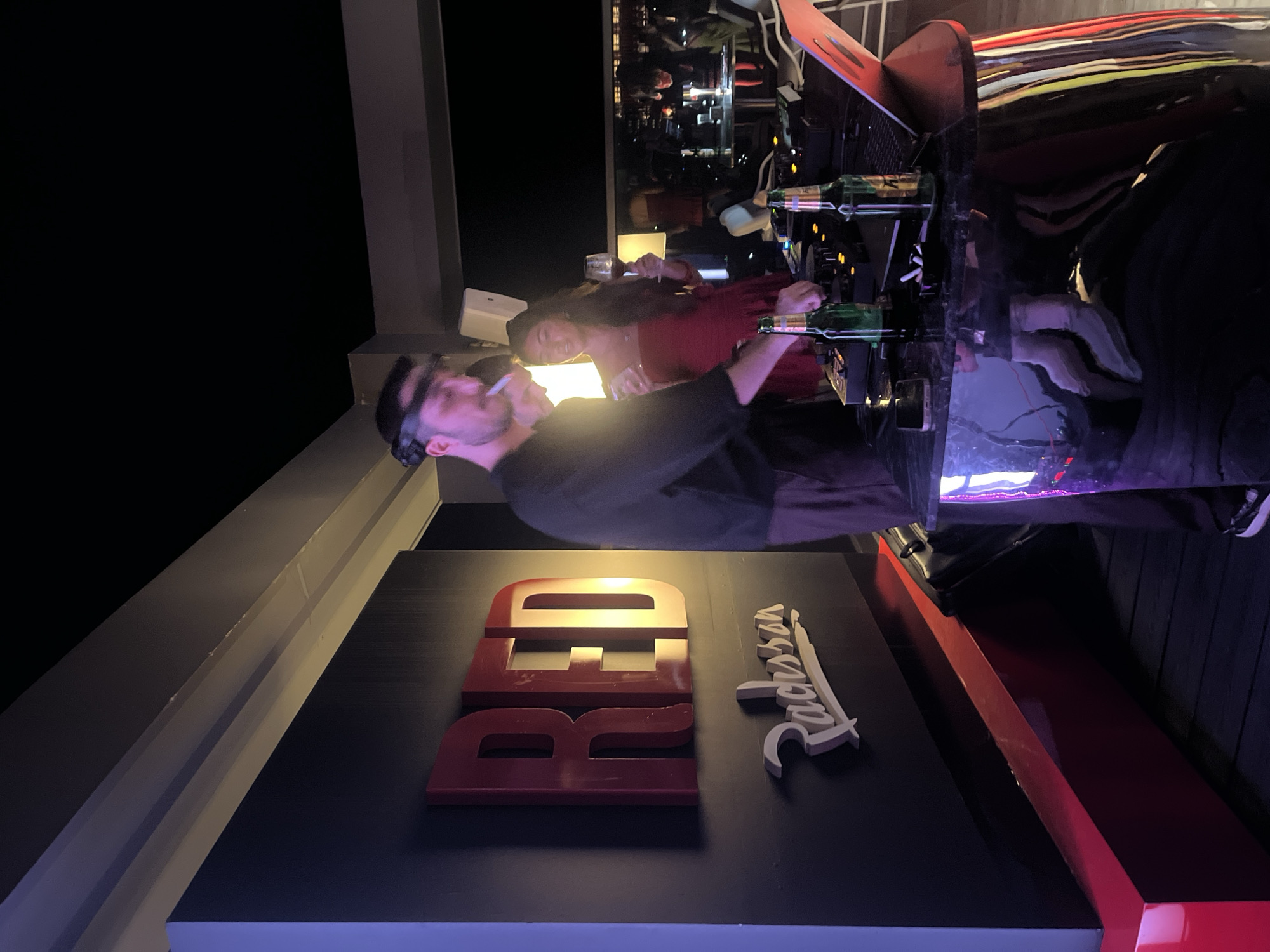 The image shows a group of people at a bar, possibly a club or a rooftop venue, at night. The scene is dimly lit, with purple and white lights casting a mood. A man in the foreground is wearing headphones and smoking a cigarette. He appears to be DJing or controlling music equipment. Behind him, a woman in a red dress is also interacting with the equipment.
In the background, other people are visible, enjoying themselves. The bar area is stocked with bottles. A prominent sign with the word "RED" in large, red, 3D letters is visible on a black surface. To the right of the "RED" sign is a white, stylized logo that appears to read "Radisson". The overall atmosphere suggests a lively social gathering. The location is identified as Đà Nẵng, Vietnam, implying an urban setting, likely an entertainment district or a hotel bar. The time of day is clearly night.