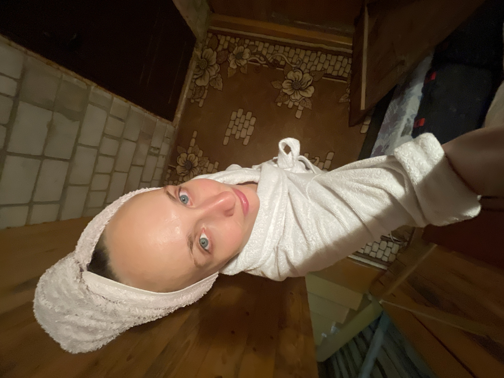 A woman with blue eyes and fair skin, her hair wrapped in a white towel, is looking up at the camera from what appears to be a sauna or bathhouse. She is wearing a white bathrobe. The background shows wooden walls, a tiled ceiling, and a patterned rug on the floor. The overall impression is that of a personal moment of relaxation. The location is described as the city of Sharkan, Russia, though this specific setting within the city is not identifiable from the image alone.