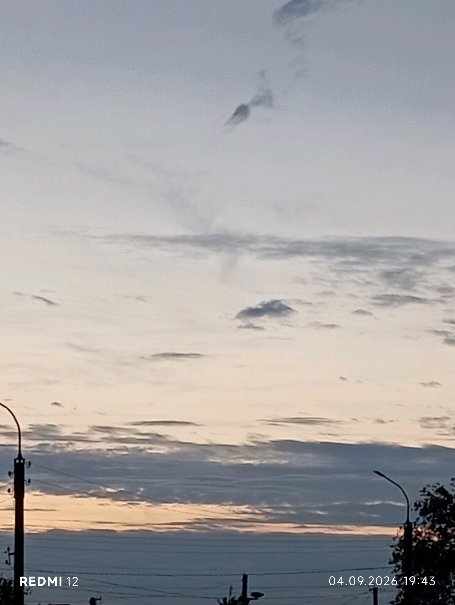 An outdoor, low-angle, full shot captures the sky above Smila, Ukraine, at dusk on September 4, 2026, at 19:43. Wispy clouds streak across a pale sky transitioning from soft yellow at the horizon to a muted gray overhead. A faint plume of smoke or vapor extends from a cloud formation in the upper portion of the frame. In the foreground, the tops of utility poles and a silhouette of trees are visible against the twilight sky. The "REDMI 12" watermark is present in the lower-left corner, and the date and time stamp "04.09.2026 19:43" is in the lower-right.
