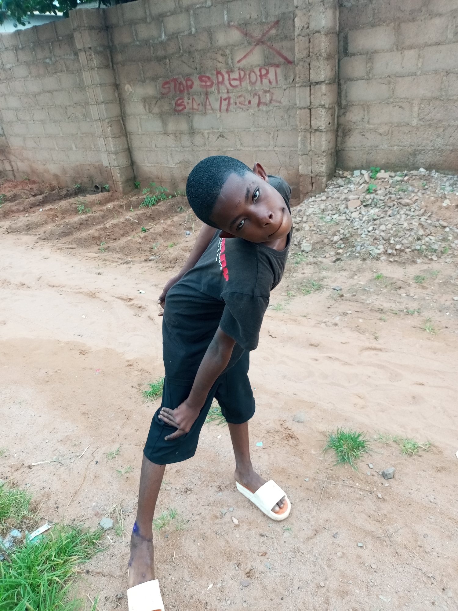 A young Black boy is standing outdoors, posing for a photo. He is wearing a black t-shirt and black shorts, and white slide sandals. He is standing with his body angled to his left, and his head tilted back and to his right, looking at the camera with a slight smile. His left hand is on his left hip, and his right arm is bent, with his hand resting on his right knee. The background is a brick wall with red graffiti. The ground is dirt and gravel. The image appears to be taken in Tempure, Nigeria.