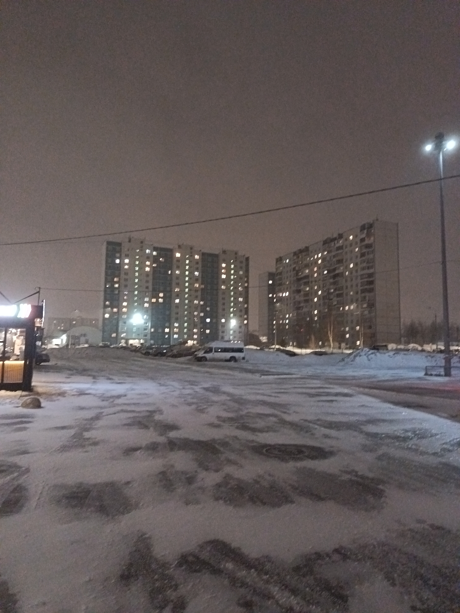The image shows a winter scene in Nizhnevartovsk, Russia, at night. Two large apartment buildings with lit windows dominate the background. In the foreground, there is a snow-covered parking lot with dark patches of exposed pavement. A streetlight is visible on the right side of the image, and a small building or kiosk is on the left. Several parked vehicles are visible in the distance. The sky is overcast, contributing to the overall dim lighting of the scene. There are no people visible.