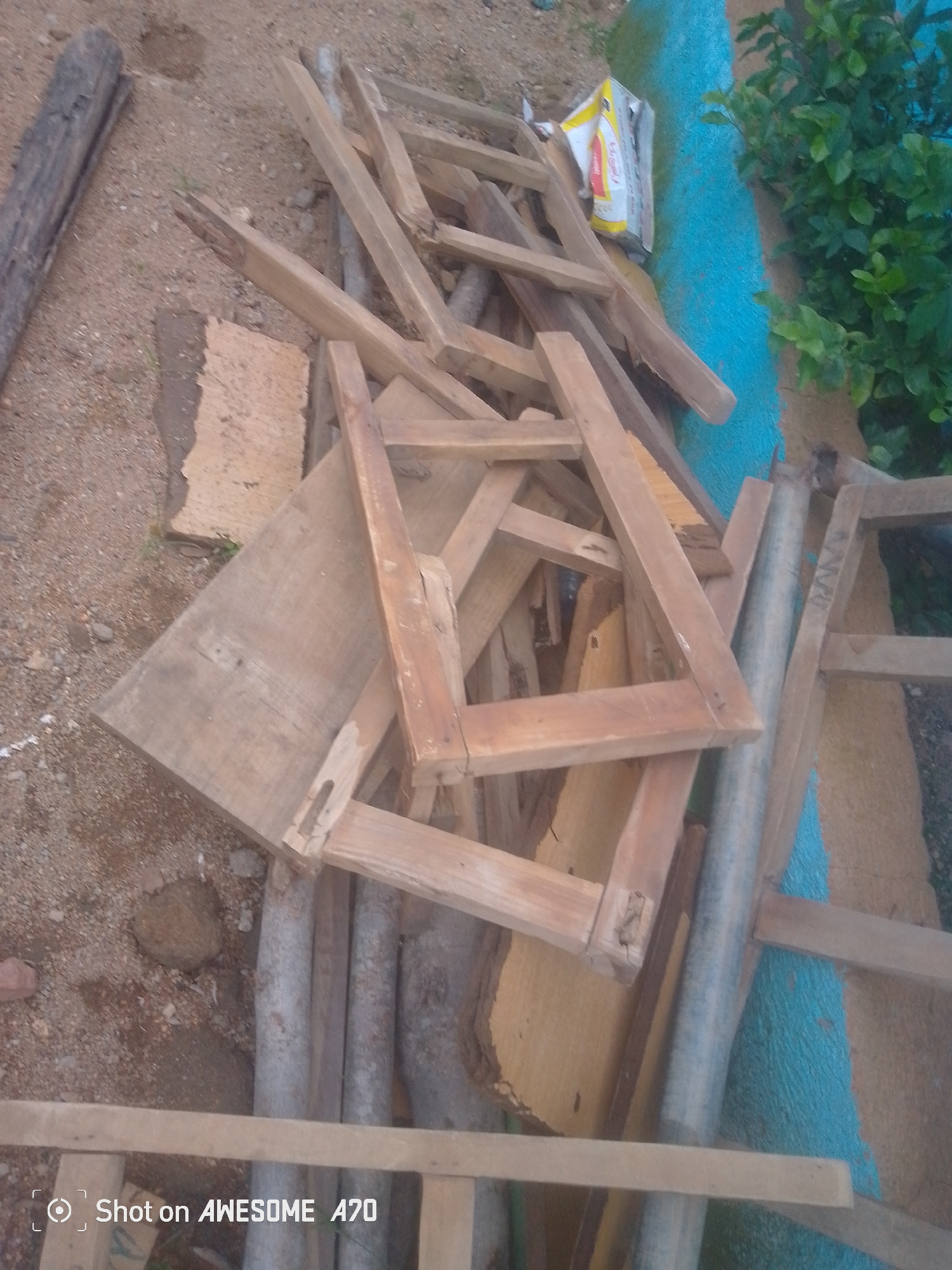 The image shows a pile of wooden planks and frames on a dirt ground, next to a blue painted wall and some green foliage. There are also some metal pipes visible. The lighting suggests it is daytime. A part of a log is visible on the left side of the image. A white and yellow bag is partially visible in the upper right quadrant of the image. There is text in the lower left corner of the image that reads "Shot on AWESOME A70".