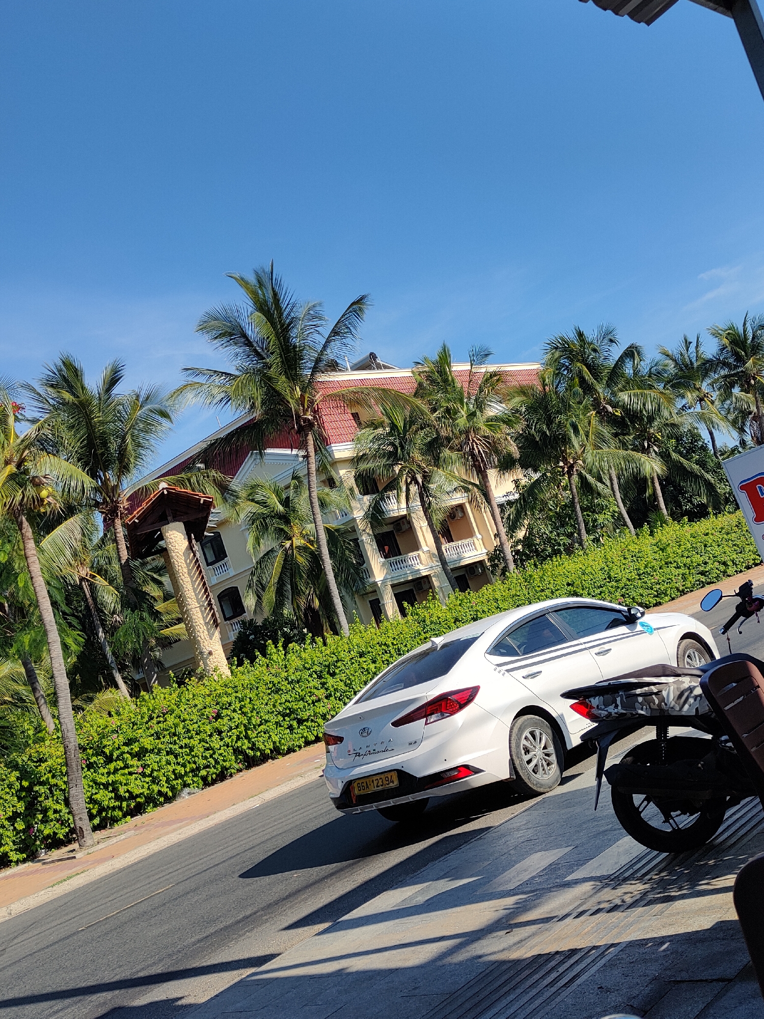 In Phu Thuy Ward, Vietnam, a white Hyundai Elantra, license plate 86A 123.94, is parked on the right side of a paved road. The car has a dark stripe on its side. In front of the car, a motorcycle with a patterned seat and a black rear fender is parked. The road is bordered by a sidewalk and a dense green hedge. Behind the hedge, a multi-story building with a red-tiled roof and cream-colored walls is visible, surrounded by numerous palm trees. The sky is clear and blue.