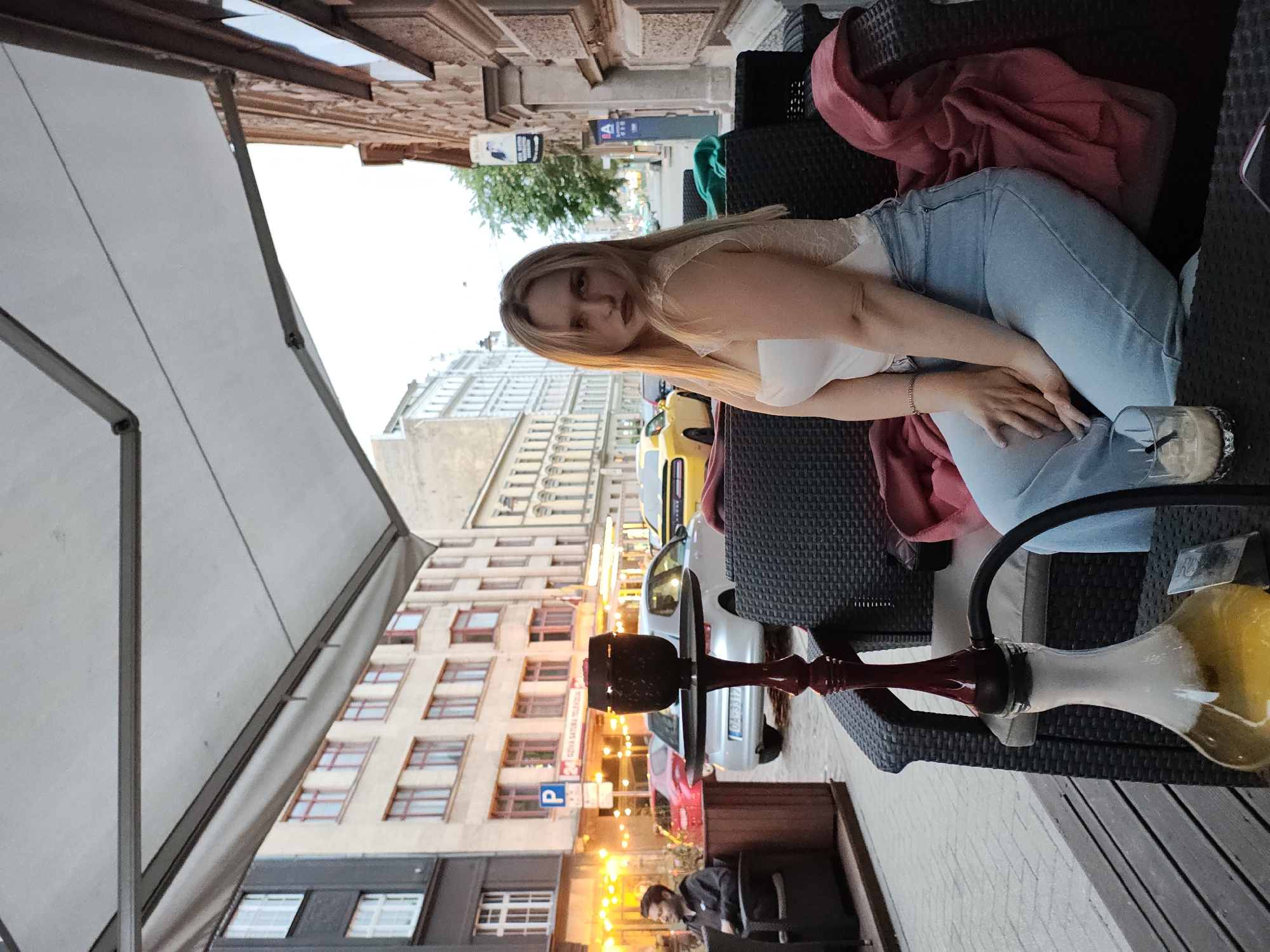 A young woman with blonde hair and dark lipstick sits on a dark wicker sofa in an outdoor seating area, facing the viewer. She wears a white top with lace detailing and light blue jeans. Her hands are clasped in her lap. To her left, a hookah with a red stem and a bulbous glass base filled with yellow liquid and smoke sits on a table. Behind her, a street scene unfolds with buildings, cars, and streetlights visible. A man in a dark jacket sits on another dark wicker chair to the far left of the frame. The sky is overcast, suggesting it is dusk or an evening setting.