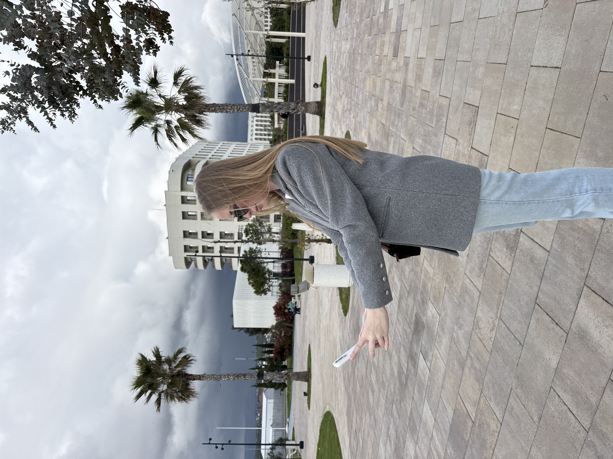 A young woman stands on a paved walkway, holding a white object in her right hand, possibly a phone or a case. She wears a grey jacket, light blue jeans, and sunglasses. Her long blonde hair hangs down her back. To her left is a multi-story white building under a cloudy sky. Palm trees and other greenery are visible near the building. A modern structure with a white roof is visible in the background, hinting at the urban setting of Sirius, Russia. The walkway is made of light-colored rectangular paving stones.