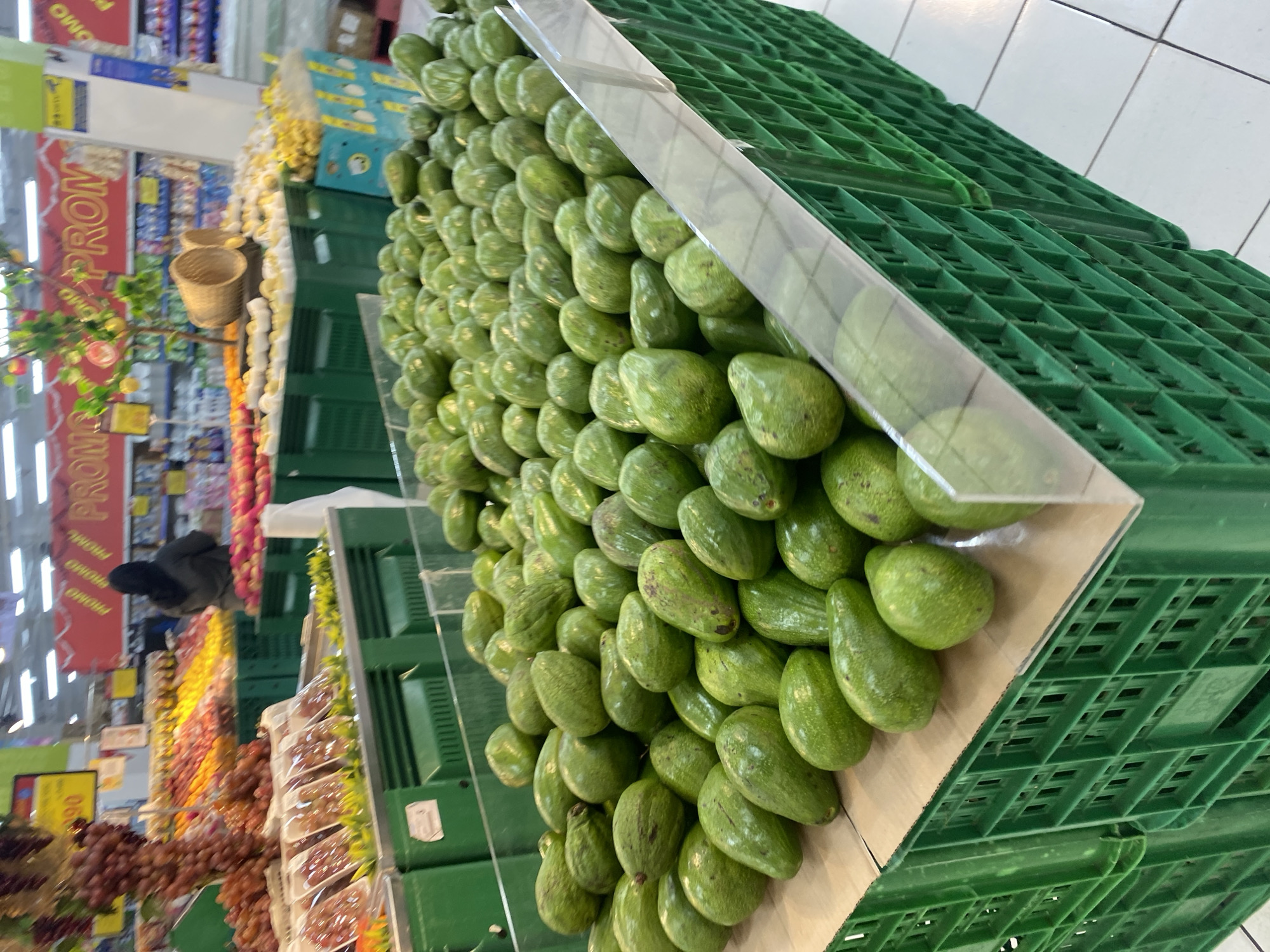 In the fresh produce section of a supermarket in Banjarbaru, Indonesia, a large display of avocados is prominently featured in green plastic crates. The avocados are a vibrant green, with some exhibiting slight blemishes or variations in color. They are piled high, filling the crates and spilling over the edges. The crates themselves are stacked and arranged in a manner that suggests a well-organized retail display. In the background, other fruits and vegetables are visible, including grapes and possibly apples, along with promotional signage. A person wearing a hijab is partially visible in the mid-ground, suggesting ongoing customer activity. The overall environment appears to be a brightly lit indoor market or grocery store.