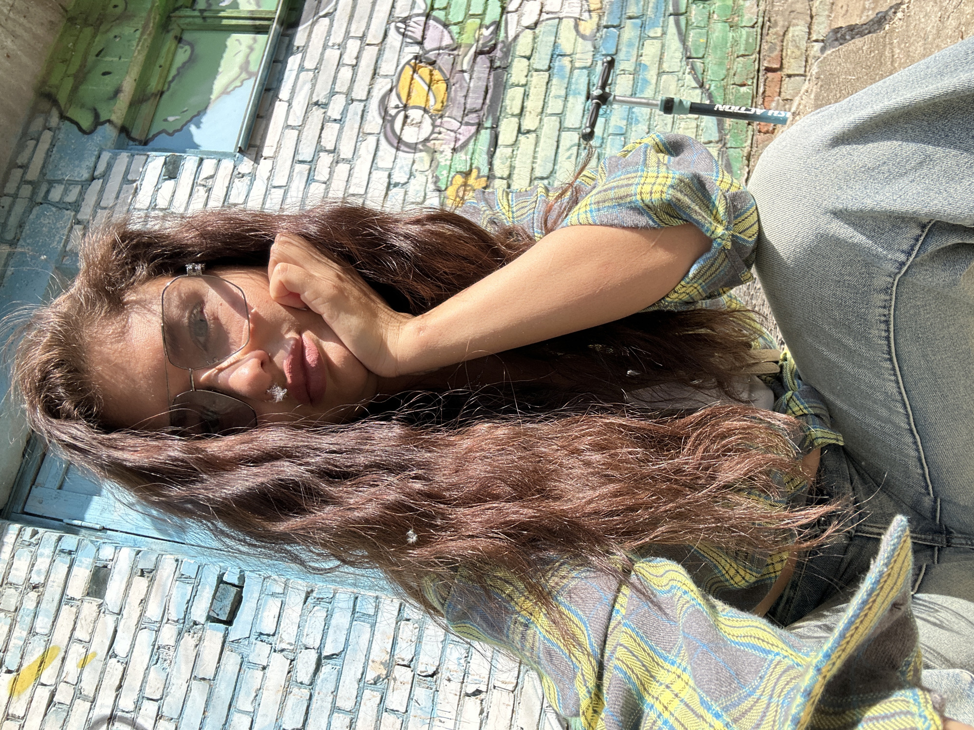 A young woman lies on her side on a painted brick surface, likely an urban wall, possibly in Khimki, Russia, based on contextual information. The woman has long, brown, wavy hair and wears wire-rimmed glasses. She wears a plaid, button-down shirt, and jeans. Her face is visible, with one hand supporting her head. The background includes a painted brick wall with both geometric and graffiti-style elements. A portion of what appears to be a scooter handlebar is visible in the background.