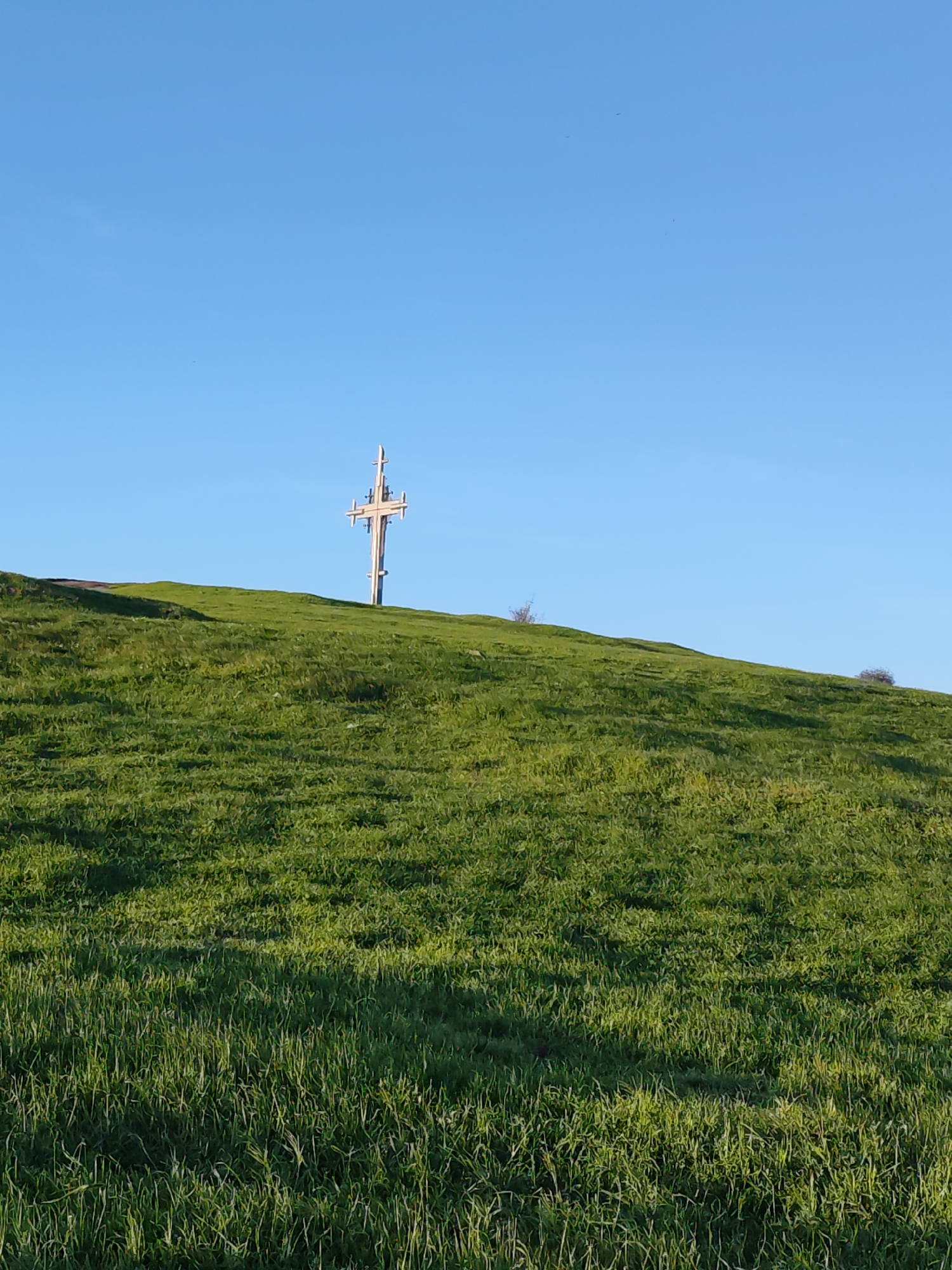 The image displays a serene outdoor landscape, primarily composed of a grassy hill and a clear blue sky. The main subject is a large, multi-armed wooden cross, likely a Calvary or memorial, positioned prominently atop the hill, slightly to the right of the frame's center. This cross is light-colored, possibly made of pale wood or painted, and features a complex structure with several horizontal beams.
The hill itself is covered in lush, vibrant green grass, indicating a healthy natural environment. Patches of darker green grass suggest variations in density or areas of shadow, hinting at the sun's position. The lower portion of the hill exhibits intricate details of individual grass blades. Small, sparse shrubs are visible on the hill's slopes, adding minor textural elements to the landscape.
The sky is a uniform, brilliant blue, completely cloudless, occupying the upper third of the photograph. The clear sky and the illumination on the cross and grass suggest bright, sunny weather. The time of day appears to be either late morning or early afternoon, given the bright, direct sunlight.
No people or significant activity are visible in the image, emphasizing the natural tranquility of the scene. The setting evokes a sense of peace and contemplation, with the cross acting as a solemn focal point against the natural backdrop. Given the context of Burgas, Bulgaria, this monument likely holds cultural or religious significance within the local landscape. There is no visible text.