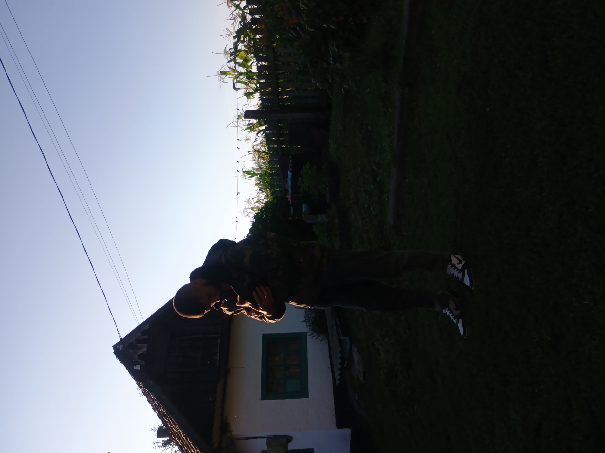 In Kliushnykivka, Ukraine, at approximately sunrise, a young man wearing a camouflage jacket and dark pants is standing in a yard, holding a small animal, possibly a cat or a rabbit, close to his chest. He is looking down at the animal. Behind him is a white house with a dark, sloped roof and a green-framed window. To the right of the house, a wooden fence encloses a garden with corn stalks reaching towards the clear, pale blue sky. The foreground is dominated by dark green grass, suggesting the area is a rural residence or yard. Power lines are visible across the sky above the house and extending towards the right of the frame.