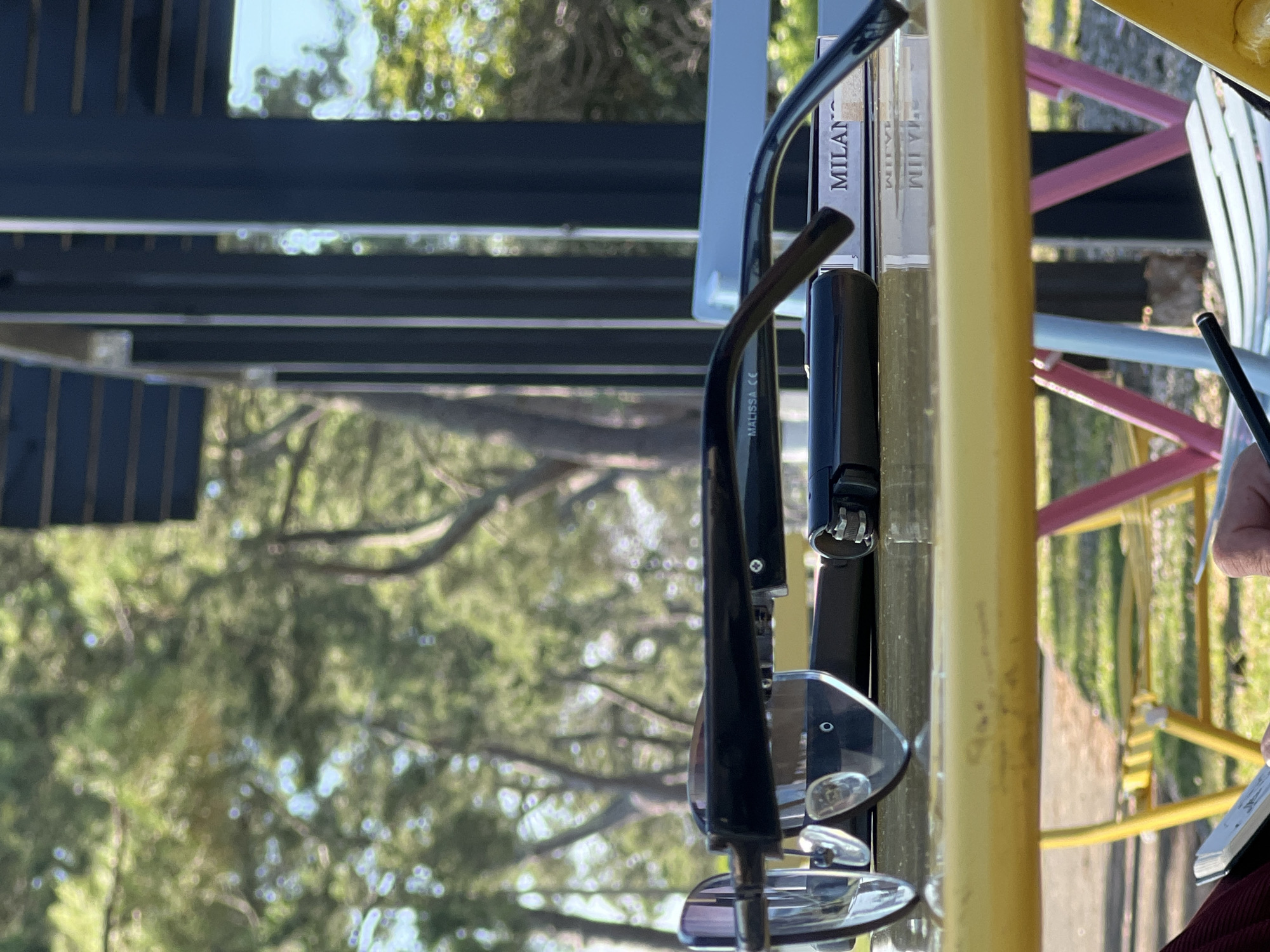 The image depicts a close-up view of sunglasses resting on a reflective surface, likely a table, in an outdoor setting. The sunglasses are black with clear lenses and are positioned in the foreground, angled to the left. Behind the sunglasses, a black cylindrical object, possibly a lighter or a pen case, is visible. Further back, a package or box with the word "MILAN" printed on it is also present. The surface they rest upon reflects the objects and the background. The background consists of blurred trees and foliage, suggesting a park or natural environment. The lighting indicates it is daytime, with shadows visible. The overall scene has a casual, relaxed feel. There is no clear indication of the city of Ochamchire, Abkhazia in the image.