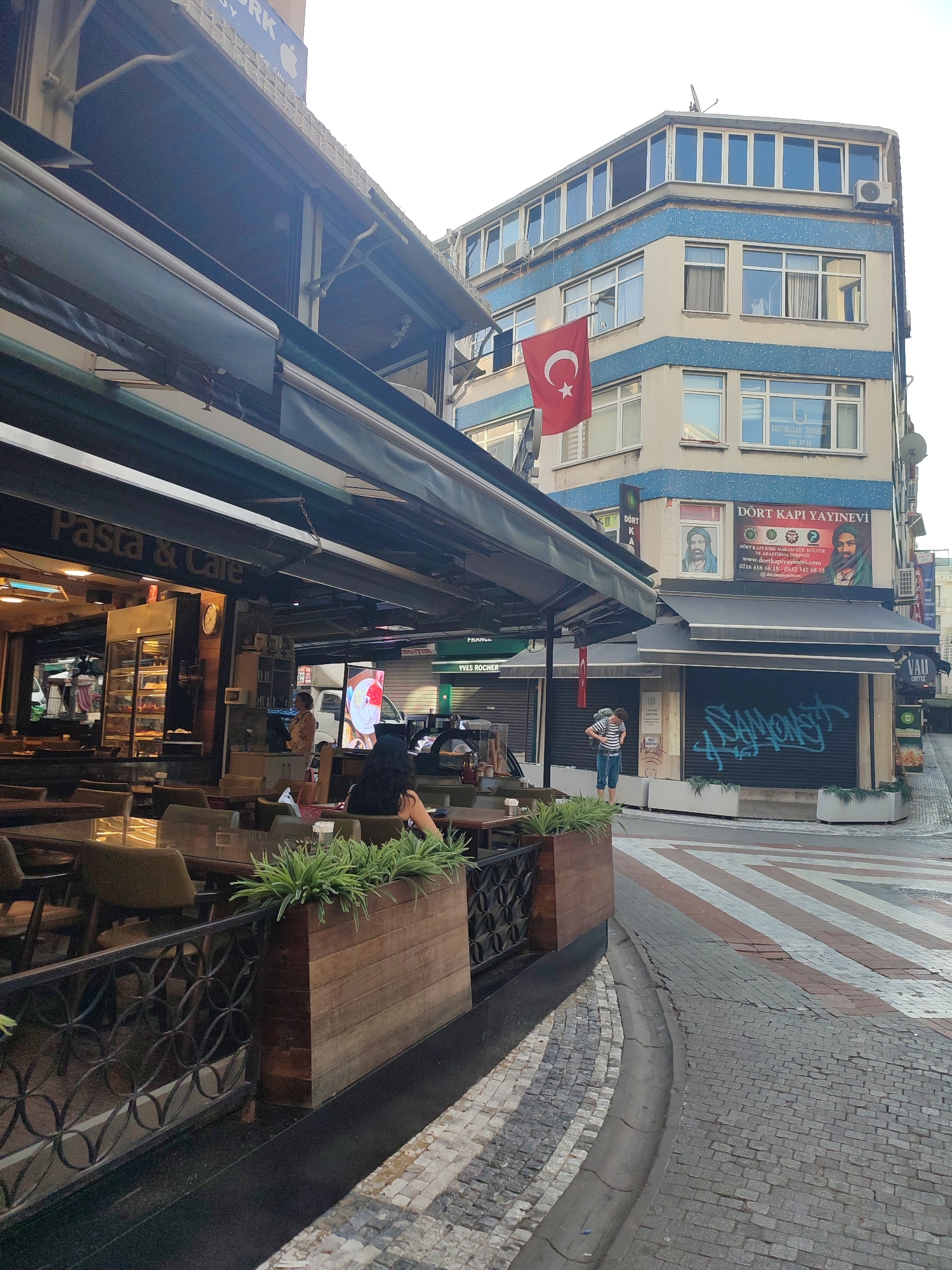 The image shows a street scene in Kadıköy, Turkey. A cafe with outdoor seating is visible on the left, with tables and chairs set up under an awning. The cafe's signage reads "Pasta & Cafe." Further down the street, several storefronts are closed, with metal shutters pulled down. One shutter is covered in graffiti. A pedestrian, wearing a striped shirt and jeans, stands on the sidewalk, facing away from the camera. A Turkish flag hangs from the side of a building on the right. The building also features windows and an air conditioning unit. The street is paved with cobblestones and has a distinct striped pattern for a crosswalk. The sky is overcast.