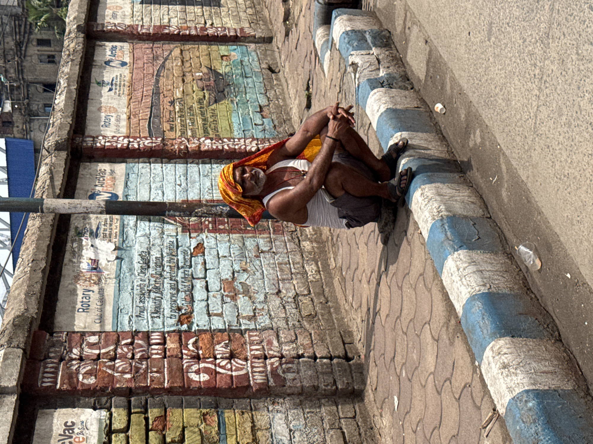 An elderly man wearing a white tank top, grey shorts, and a yellow and orange patterned head covering sits cross-legged on the edge of a paved walkway in Kolkata, India. He rests against a painted brick wall featuring a Rotary Club advertisement reading "Proud Asicions of Clean Ganga & Beautify Kolkata Joiitiative to preserve and protect the Heritage Sultur of Kolkata riverside." The wall also displays the logos of "Arvac" and "Rotary" alongside the Rotary Club of Calcutta's name. The walkway border is painted with alternating blue and white stripes. A few pieces of litter are scattered on the ground near the border. A grey pole stands to the left of the man, and there's a partial view of a building in the upper left corner of the frame.