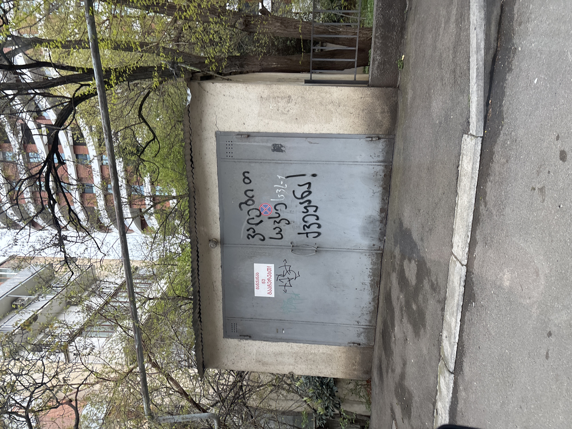 An outdoor scene in Tbilisi, Georgia, features a concrete storage unit or garage with a grey, weathered double metal door. The structure has a slightly angled corrugated roof. On the left side of the image, bare tree branches with emerging green leaves partially obscure modern apartment buildings in the background. A thin, vertical pole is visible in the mid-ground. To the right of the garage, there's an asphalt road or pathway with a concrete curb.
The main focus is the graffiti on the garage doors. On the left door, large black letters spell out "კაცია თუ" (Is it a man or) followed by a stylized red and white symbol that resembles a "no parking" sign (a crossed-out 'P' within a circle), but uniquely incorporates a heart shape and some Georgian letters. After the symbol, the text continues "ქალია" (or a woman), then below that, "63-1". On the right door, in large black letters, is the word "წავედი!" (I'm gone! / I left!).
Below the main graffiti on the left door, a smaller red and white sticker or sign reads "გასაღები 63-1" (Key 63-1) and underneath that, "გაჭედავთ!" (You will jam it! / You will block it!). There are also some faded, less discernible scribbles and tags on the lower parts of the doors.
The overall impression is daytime in spring, given the fresh foliage. There are no people visible, and the scene is static, capturing an urban setting marked by graffiti and practical signage.