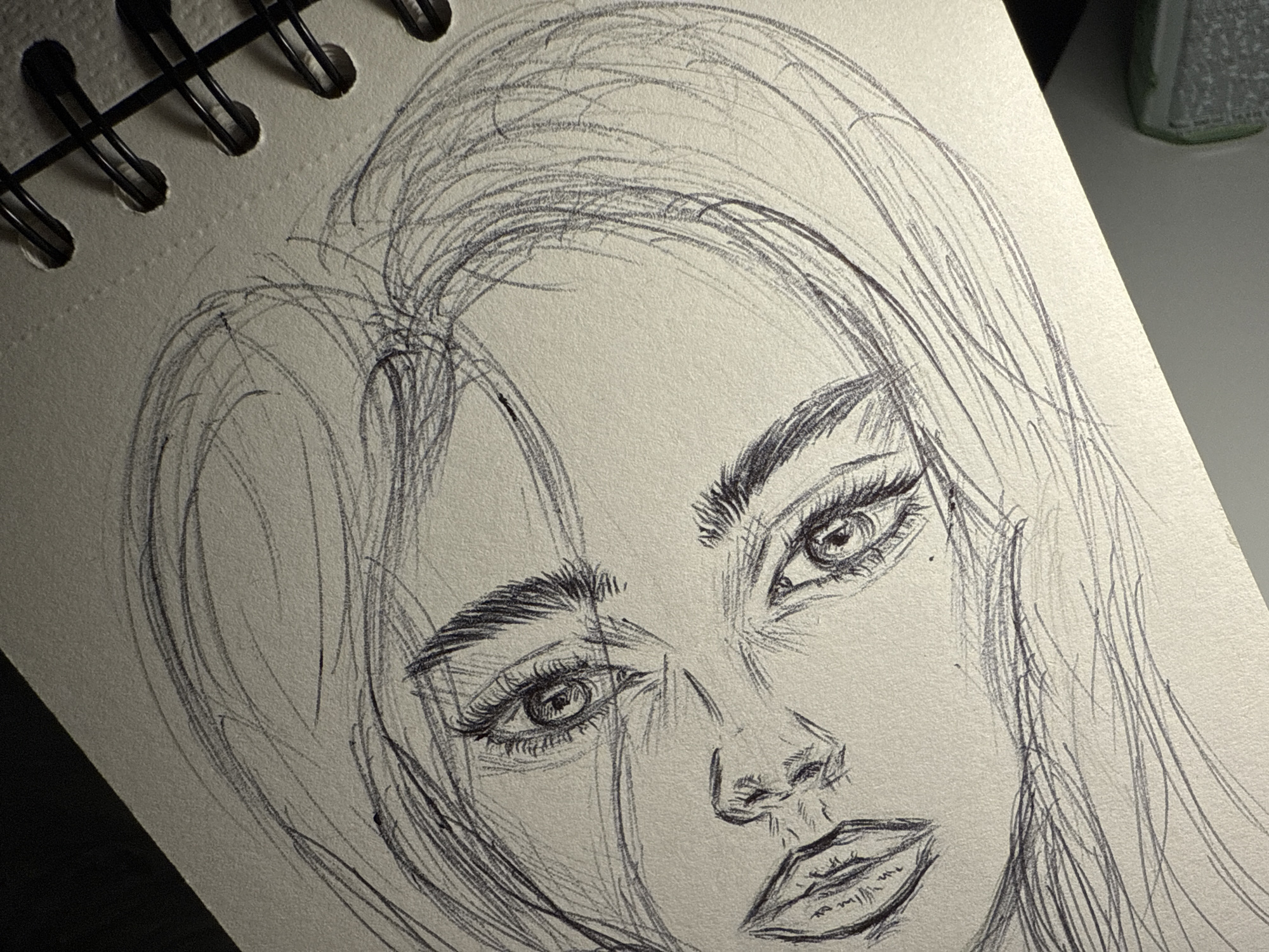 A close-up sketch of a woman's face is drawn on a white notebook page, which is bound by black metal rings at the top left corner. The sketch is done in pencil, showing detailed features like eyes, eyebrows, nose, and lips. The hair is loosely drawn, flowing around the face. The notebook sits at an angle. In the upper right corner is a small, out-of-focus object, possibly a green container or eraser, partially obscuring the background. No specific location in Sofia, Bulgaria is identifiable.