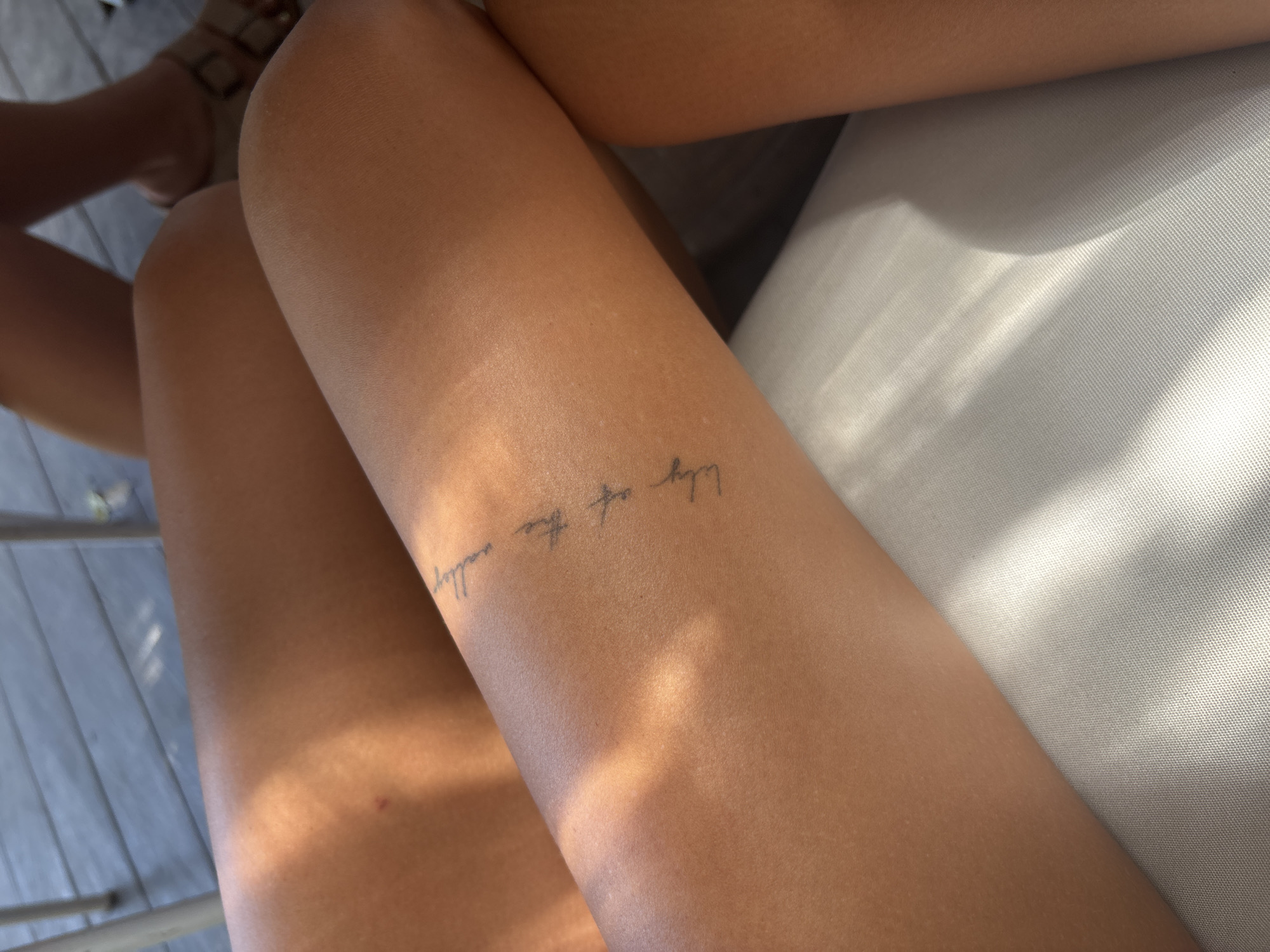 The image shows a close-up of a person's legs. The right leg has a tattoo on the upper thigh area that says "Lily of the valley." The right leg is resting on a white woven surface, possibly a cushion or chair. Another leg is visible on the left side of the image, partially cropped out. The background includes a partially visible wooden deck or floor, and the top of a foot wearing a sandal. The location is Kuta Selatan, Indonesia.