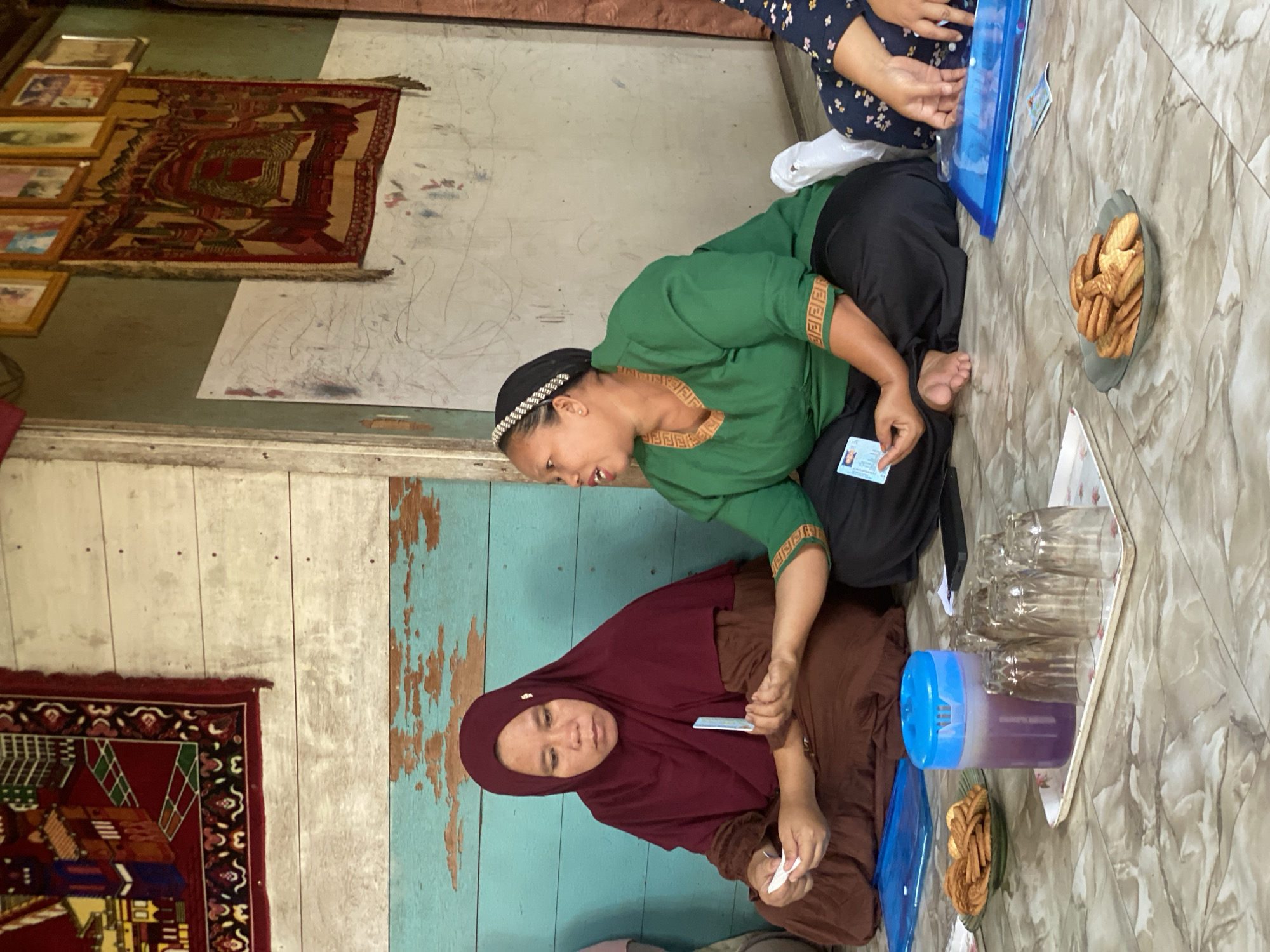 In Bawahan Selan, Indonesia, two women are seated on a marble-patterned floor. The woman on the left wears a maroon hijab and brown clothing, holding a white card. The woman on the right is dressed in a green top and black pants, wearing a beaded headband, and is holding a blue identification card. They are facing each other, appearing to be in conversation. A tray with a blue pitcher filled with purple liquid, several clear glasses, and a plate of cookies sits between them. Another plate of cookies is visible to the left. The background features a light-colored wooden wall with turquoise accents, adorned with framed pictures and two woven tapestries. Another person's legs are visible in the lower right corner, wearing a dark blue patterned skirt.