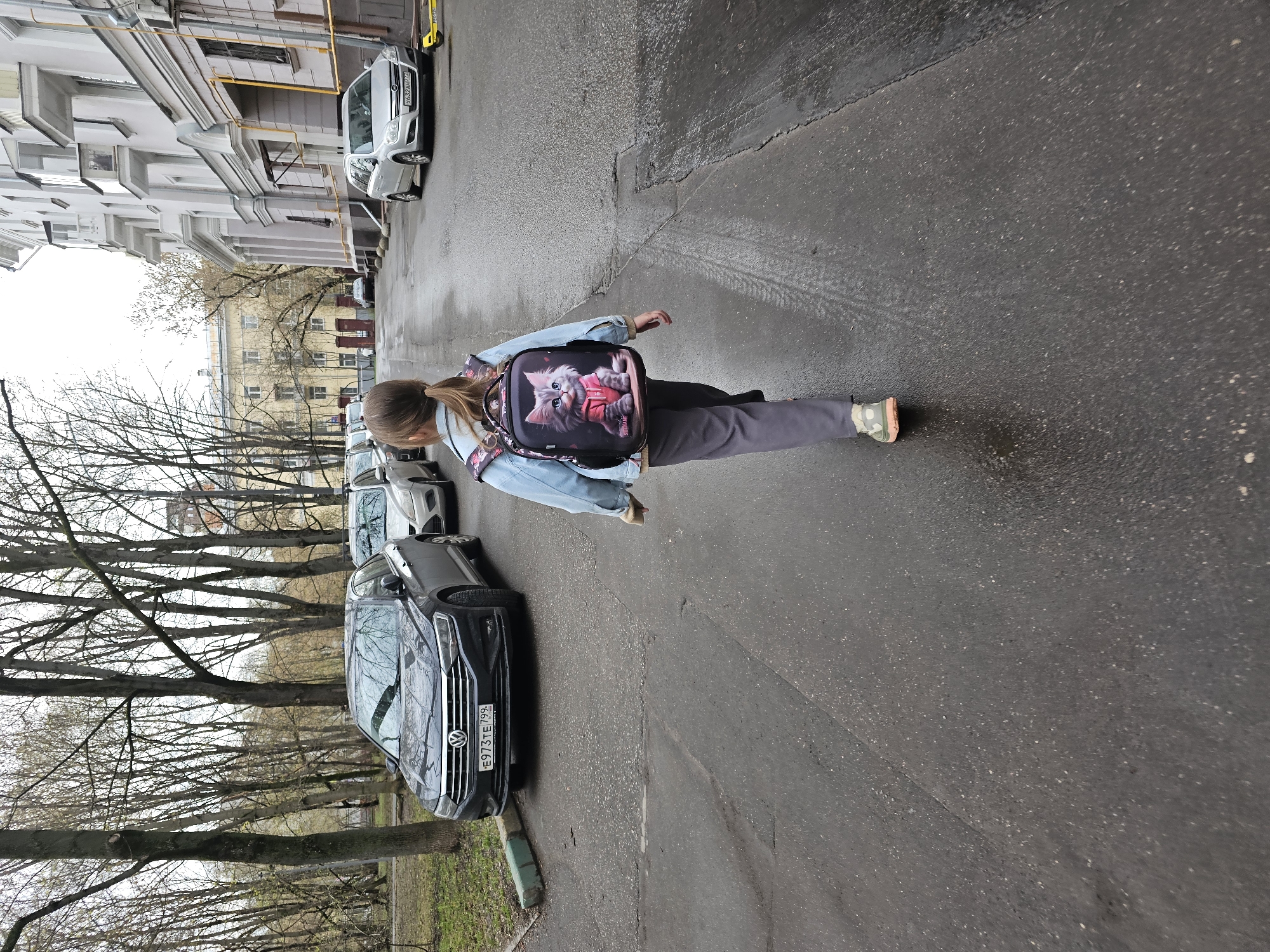 A girl with a ponytail walks on the wet asphalt on a street in Moscow, Russia. She is wearing a light blue denim jacket and carrying a backpack with a picture of a cat on it. Along the left side of the road, bare trees line a patch of grass, with parked cars behind them. The black car closest to the camera has the license plate "E973 TE 799". Tall buildings are visible in the background, and parked cars line the right side of the road.