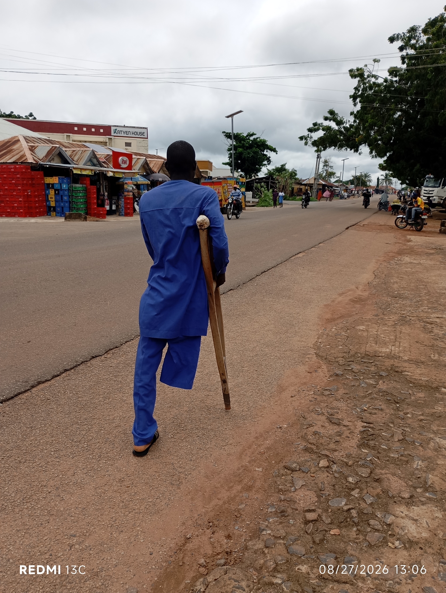 A man with an amputated left leg, using crutches, walks along the side of a road in Shendam, Nigeria. He is wearing a blue kaftan and trousers. In the background, there are shops with red and white awnings, crates of beverages, and a sign that reads "KATYEN HOUSE." The road is paved and appears to be a busy thoroughfare with several motorcycles and a truck visible in the distance. The sky is overcast, suggesting a cloudy day. The scene is captured from a low angle, looking from behind the man.