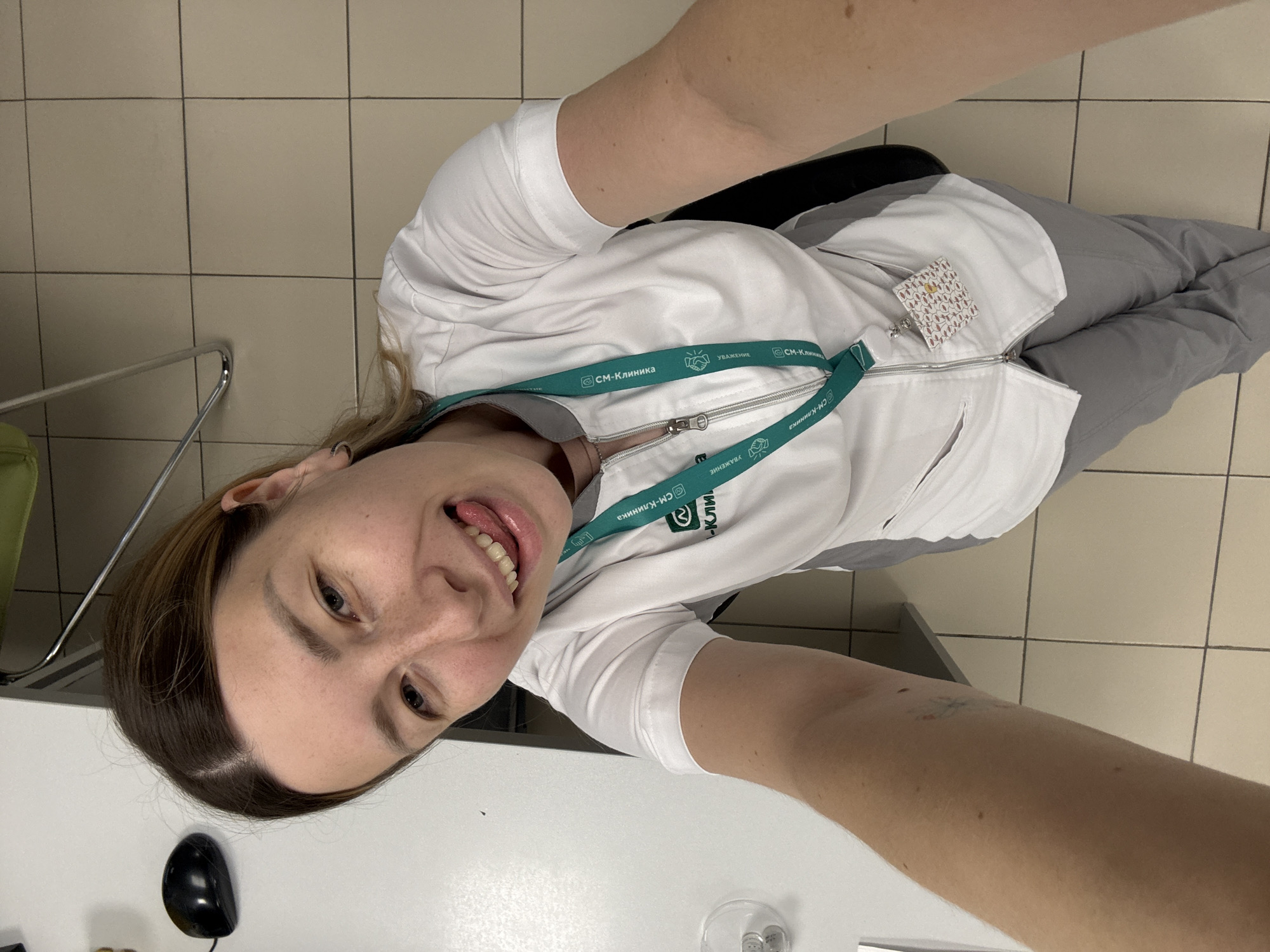 A woman is taking a selfie in a tiled room. She is wearing a white medical scrub top with grey pants and a lanyard with the logo of "SM-Clinic" around her neck. The woman has light brown hair and is sticking her tongue out at the camera, smiling. To her left on a white desk, there is a black computer mouse. The floor is tiled with cream-colored squares. The background includes a metal chair and a sliver of a green chair. The scene appears to be indoors, possibly in a clinic or hospital setting, given the uniform and the logo. The lighting is bright and appears to be overhead. The overall mood of the image is playful and informal.