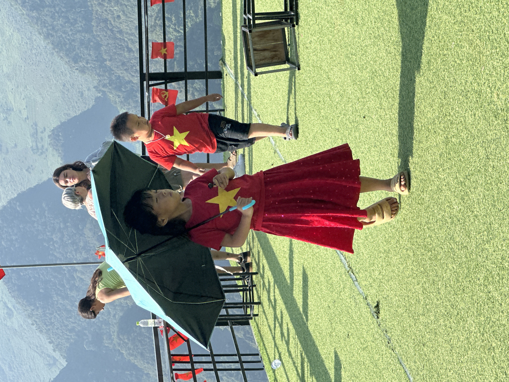A view from above shows two children and several adults on a grassy, artificial surface in Đường Thượng Commune, Vietnam. The children, a boy and a girl, are both wearing red shirts with a yellow star, the symbol of Vietnam. The girl is also wearing a red dress. She is holding a dark umbrella with blue trim. Vietnamese flags are visible attached to the railing. A mountainous landscape forms the backdrop.