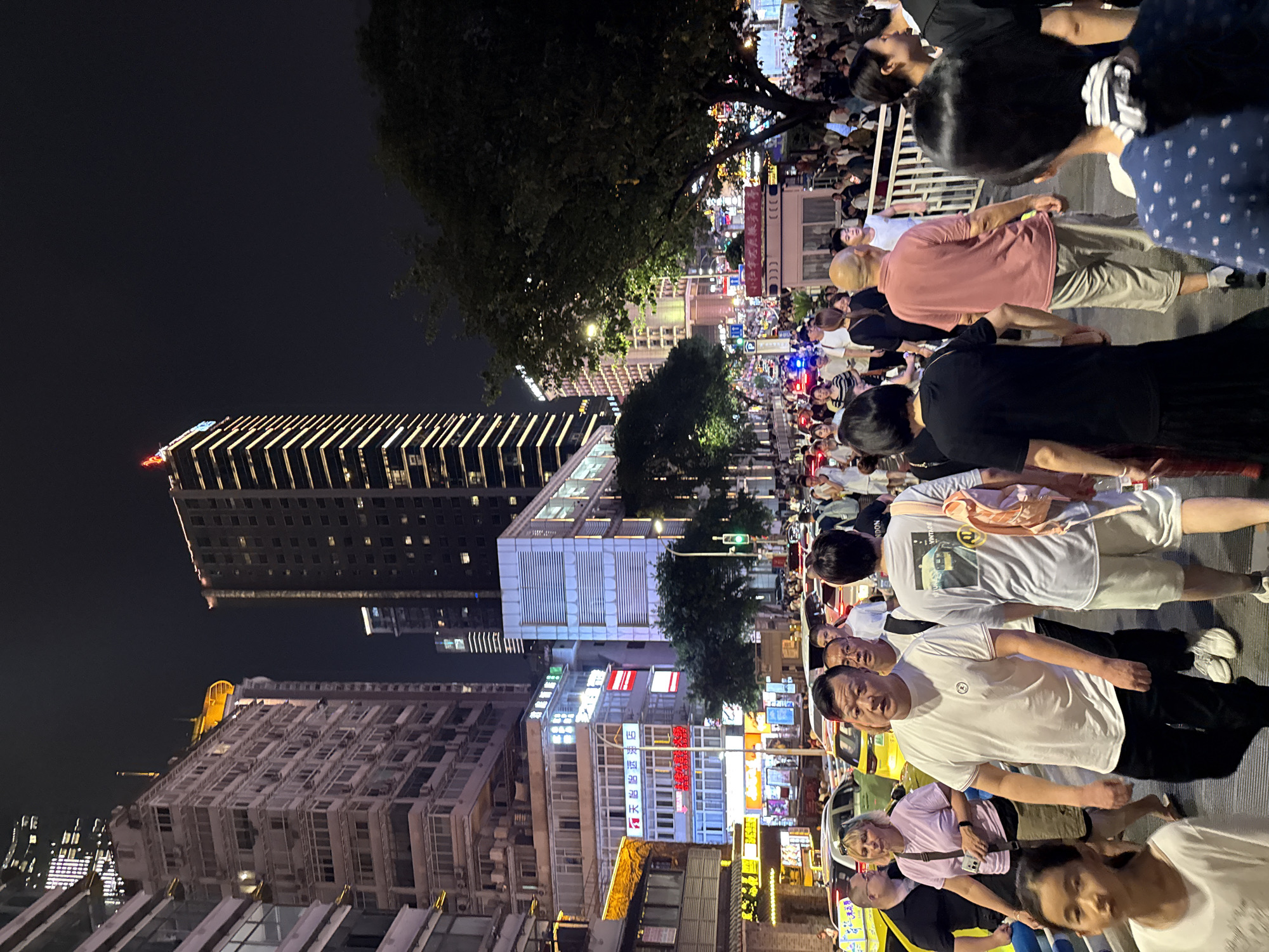 The image captures a bustling nighttime street scene in Yuzhong District, China, likely a commercial or entertainment area given the numerous buildings with illuminated windows and signage. A dense crowd of people is walking on a paved pathway alongside a street filled with vehicles, including cars and what appear to be taxis. The architecture is dominated by tall, modern buildings, some featuring distinctive lighting designs. Trees are visible, interspersed with the urban structures. The overall atmosphere is lively, suggesting a popular gathering spot or a busy thoroughfare.