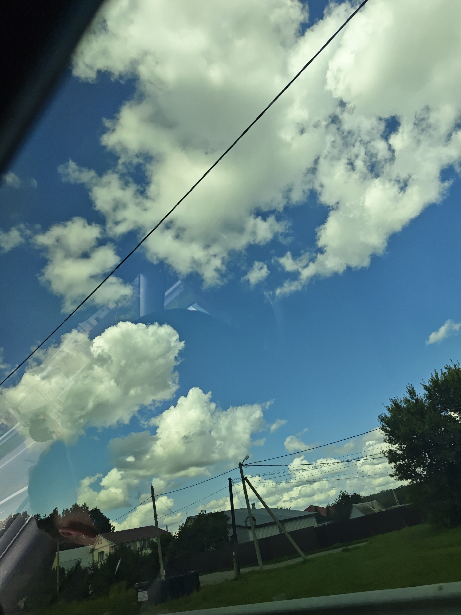 The image captures a view of a rural landscape under a bright blue sky with scattered white clouds. In the foreground, a dark, reflective surface, likely a car window, partially obscures the scene. A thin black wire runs diagonally across the upper portion of the frame. Below this, the sky is visible with fluffy cumulus clouds.
Further down, a residential area is depicted. Several houses with red and grey roofs are visible amidst dense green trees and foliage. A dark brown fence runs along the properties. Electrical poles with wires are interspersed throughout the scene. The ground in front of the houses is covered in green grass, and a dark object, possibly a garbage bin, is present. The overall impression is a sunny day in a quiet, rural setting.