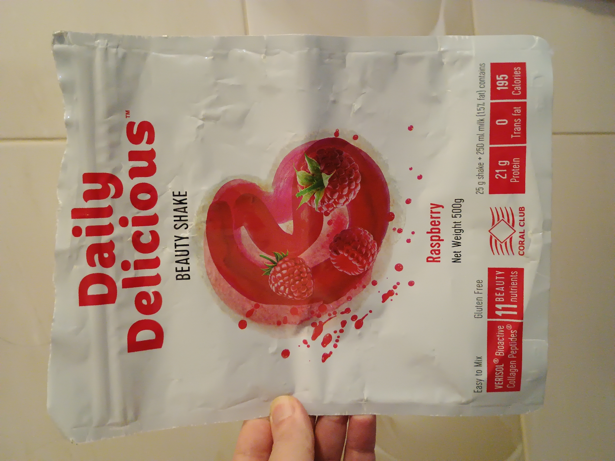 The image shows the front of a white plastic pouch of "Daily Delicious" Raspberry Beauty Shake. The pouch has a large graphic of raspberries and a swirling red liquid. Text on the pouch includes "Daily Delicious", "BEAUTY SHAKE", "Raspberry", "Net Weight 500g", and nutritional information. The pouch is held by a person's hand on the left side. The background appears to be a tiled wall. There is no information in the image to suggest its location in Zaporizhzhia, Ukraine.