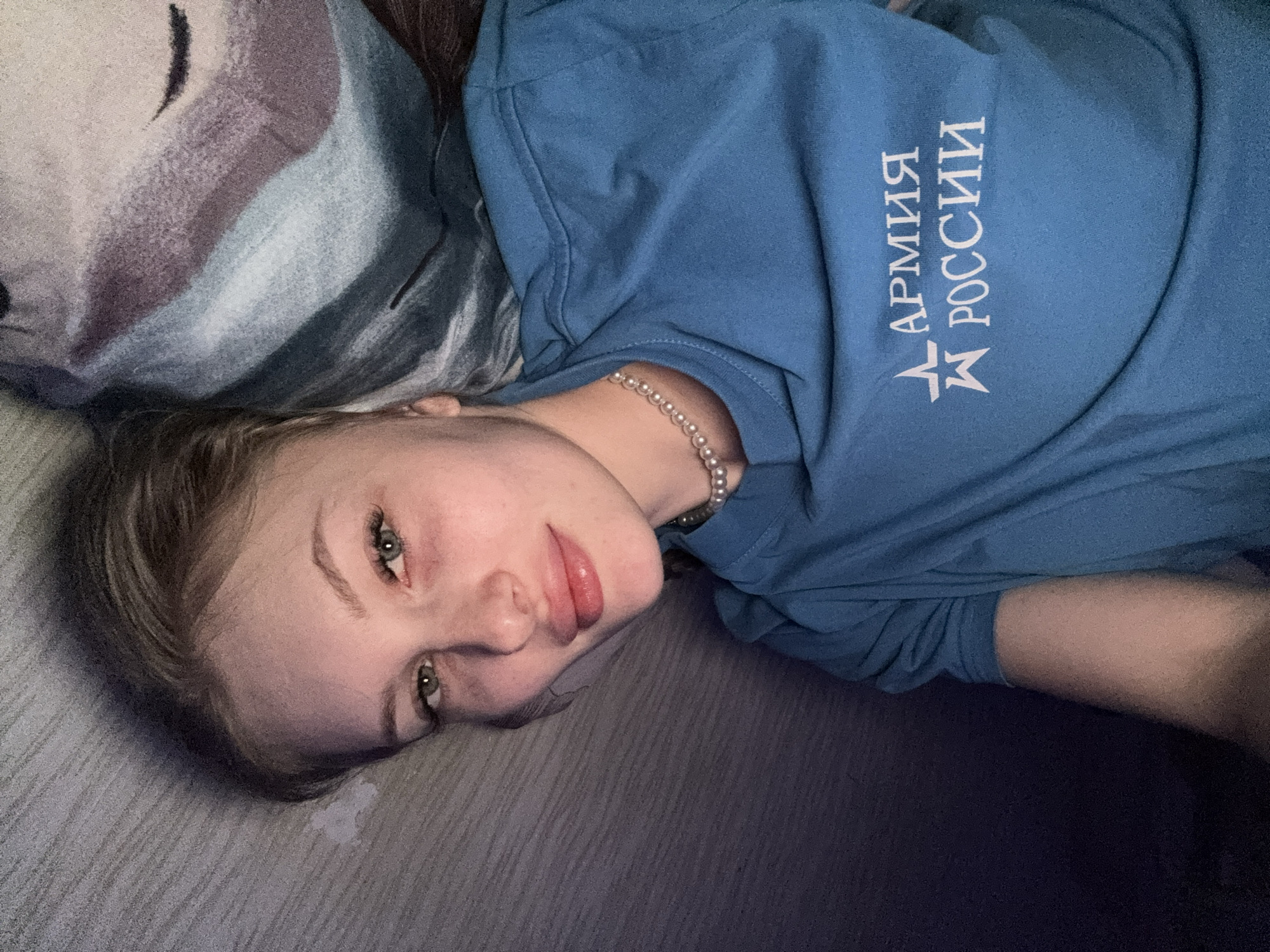 A young woman is depicted in a relaxed, close-up shot, lying on her side and gazing directly at the camera. She has fair skin, light-colored hair, and striking light eyes, possibly green or hazel, with clearly defined eyebrows. Her lips are slightly parted, showing a natural, soft pink color. She appears to have a calm and composed expression.
She is wearing a blue, long-sleeved sweatshirt or top. On the chest of her shirt, white Cyrillic text is visible, reading "АРМИЯ РОССИИ" (ARMIA ROSSII), which translates to "ARMY OF RUSSIA," alongside a stylized star emblem. A delicate pearl necklace is worn around her neck.
Her head rests on a pillow adorned with a soft, abstract pattern in shades of white, grey, and purple. The woman is situated on a dark, possibly grey, textured surface, likely a bed or couch, suggesting an indoor setting. The lighting is soft and ambient, contributing to a serene mood, perhaps indicating evening hours or an intentionally subdued environment. This casual and intimate moment takes place in Кондратово, Russia.
