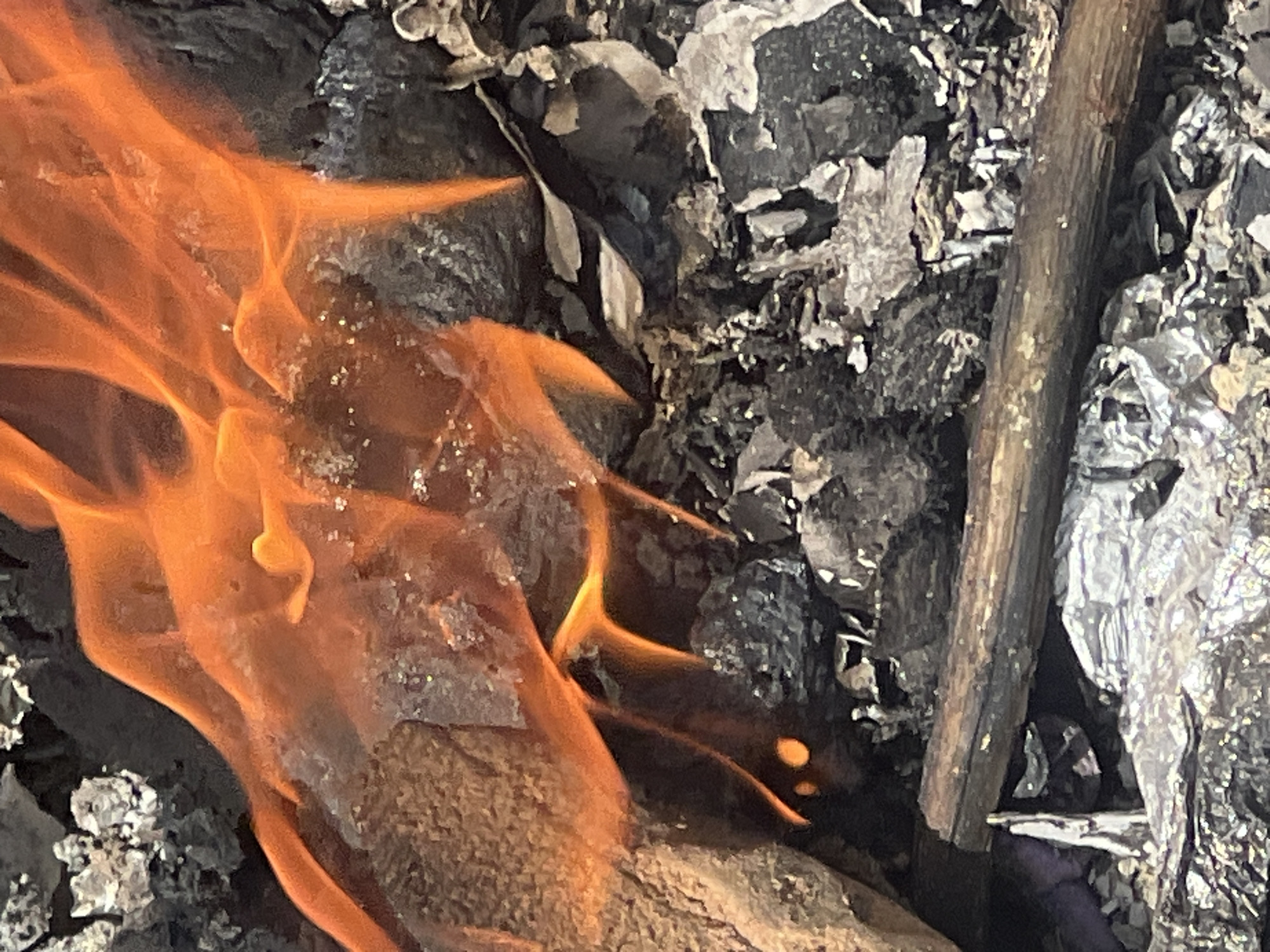 A close-up shot depicts a fire burning amidst charred wood, ash, and foil-like debris. No people are visible in the frame. The exact location within the city of Gura Top, Nigeria cannot be determined from this image alone.