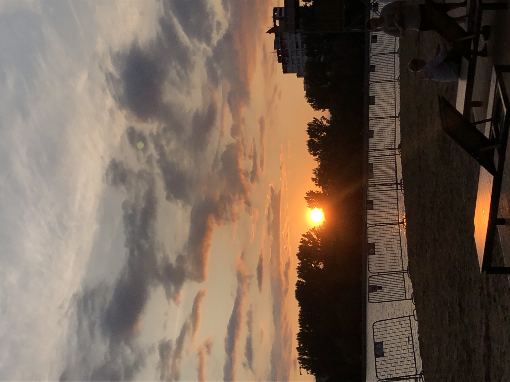 The image captures a sunset over a body of water, likely a river or lake, on a beach in Khimki, Russia. The sky is filled with dramatic clouds illuminated by the setting sun, casting a warm orange glow across the scene. Silhouetted trees line the water's edge, and a few people are visible on the sandy beach in the foreground. Two individuals stand near the water, one appearing to hold something. A young person is seated on a lounge chair, and another lounge chair is empty. Metal barriers separate the beach from the water. In the background, a multi-story building, possibly an apartment complex or hotel, is visible, along with a construction crane. The overall atmosphere is serene, with the natural beauty of the sunset juxtaposed against the urban landscape.