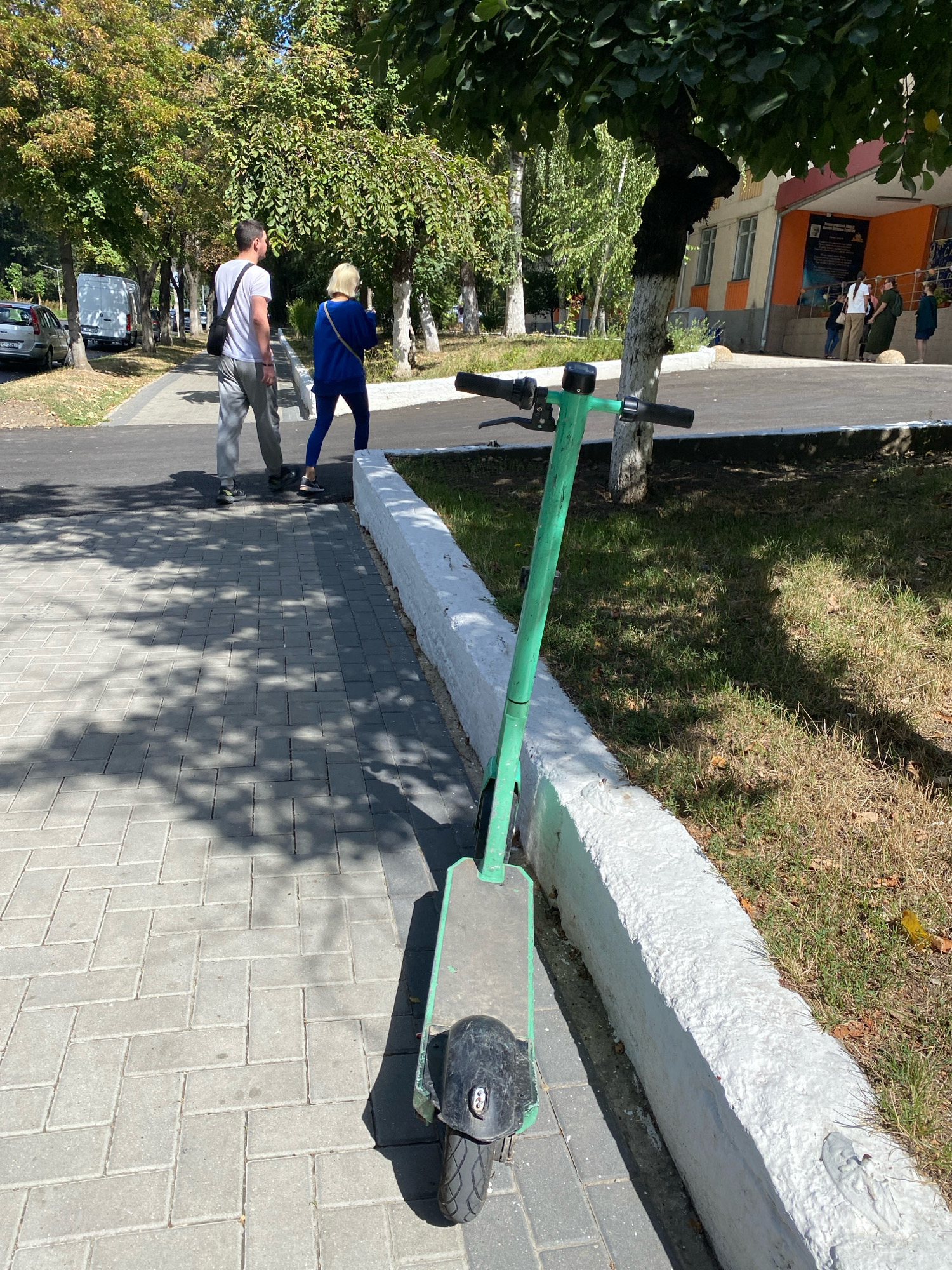 This image depicts a bright, sunny day in Chișinău, Moldova. In the foreground, a green electric scooter is parked on a grey brick sidewalk. The scooter is oriented towards the viewer, with its front wheel and handlebars clearly visible. The sidewalk is partially covered in dappled shade cast by overhead trees. A low, white concrete barrier runs along the edge of the sidewalk, separating it from a grassy area.
In the middle ground, a man and a woman are walking away from the viewer on the sidewalk. The man is wearing a white t-shirt, grey pants, and dark shoes, with a dark bag slung over his shoulder. The woman is wearing a bright blue tracksuit and dark sneakers. They are walking towards a building visible in the background.
The background features a building with an orange and beige facade, with several windows and an entrance. A few other individuals are visible near the building entrance. Mature trees with green foliage line the sidewalk and the area around the building, suggesting a park-like or campus setting. A white van is parked on the street to the left of the image. The overall scene conveys a casual, outdoor urban environment.