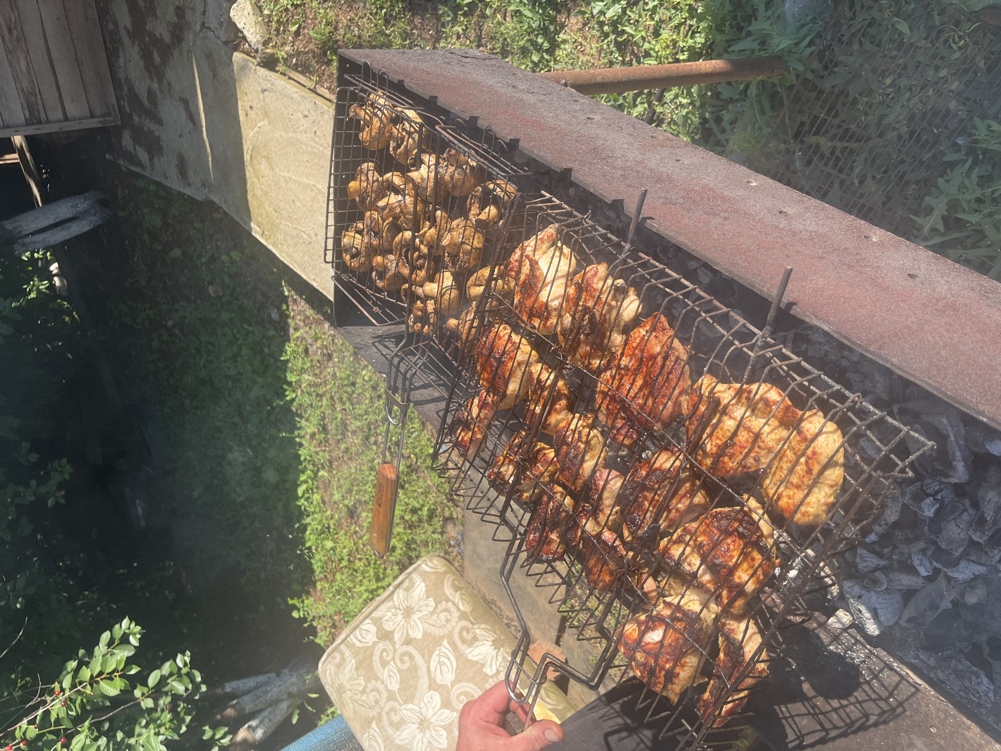This media file captures a close-up, overhead view of a barbecue with two grilling baskets filled with food. The basket on the left contains numerous whole mushrooms, lightly seasoned. The basket on the right is filled with pieces of chicken, some whole, some cut, all visibly browned and charred from grilling. The chicken pieces are arranged in multiple layers within the basket, with some appearing to be whole Cornish game hens.
The barbecue itself appears to be a rustic, metal construction, possibly homemade, resting on a bed of glowing coals. The background shows an outdoor setting with green foliage and grass, suggesting a garden or natural environment. A concrete structure is visible to the left of the barbecue, possibly a part of a patio or pathway. A wooden element, likely part of a wooden fence or structure, is also present in the upper left corner. A weathered metal pipe runs horizontally behind the barbecue.
A human hand is visible in the foreground, holding the handle of the grilling basket, indicating active preparation of the food. A patterned cushion or chair is also partially visible in the lower part of the image, adding to the casual, outdoor dining atmosphere. The lighting suggests it is daytime, with the sun casting shadows. The overall mood is relaxed and indicative of a summer outdoor meal. There is no visible text in the media file.