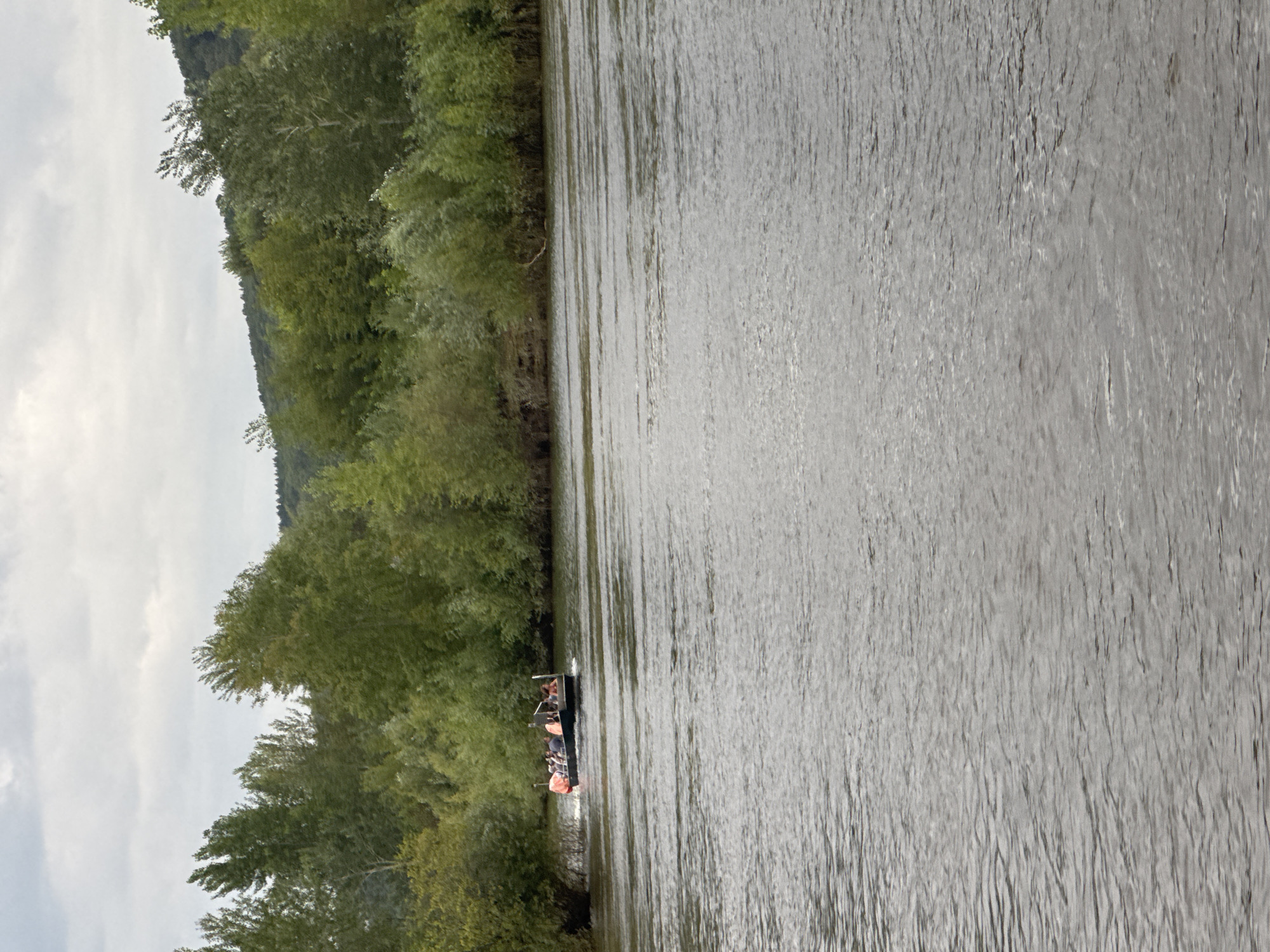 A wide, calm river flows towards the viewer under a cloudy sky. Dense green trees line the far bank, with a gentle, forested hill rising behind them. In the mid-ground, a dark, flat-bottomed boat with an outboard motor is moving to the left, carrying at least six people. Some of the passengers appear to be wearing orange life vests. The water has a rippled surface, reflecting the overcast sky. This scene is consistent with the Belaya River embankment in Ufa, Russia.