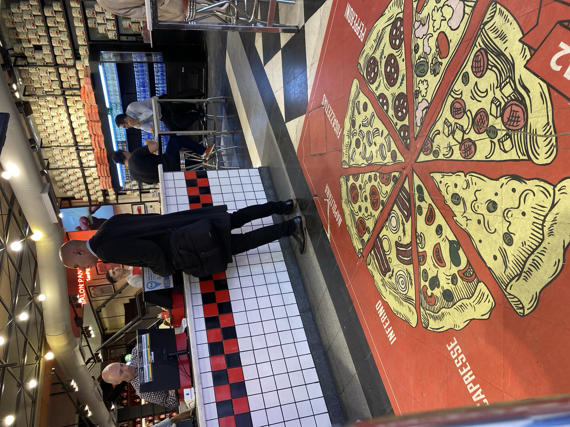 The image depicts the interior of a pizza restaurant in Buenos Aires, Argentina. A bald man in a dark suit is standing near the counter, facing away from the camera. Two other people are visible behind the counter. A cashier is working at a computer. In the background, two patrons are seated at a table. One is facing the camera, and the other is looking down. Behind them, shelves are stacked with canned goods and refrigerated drinks. The floor is tiled in a black-and-white checkerboard pattern near the counter, and transitions to a red floor featuring a cartoon-style graphic of a whole pizza cut into slices labeled with different flavor names such as "Pepperoni," "Fugazettas," "Napoletana," "Inferno," and "Capresse". The overall lighting is bright from overhead fixtures.