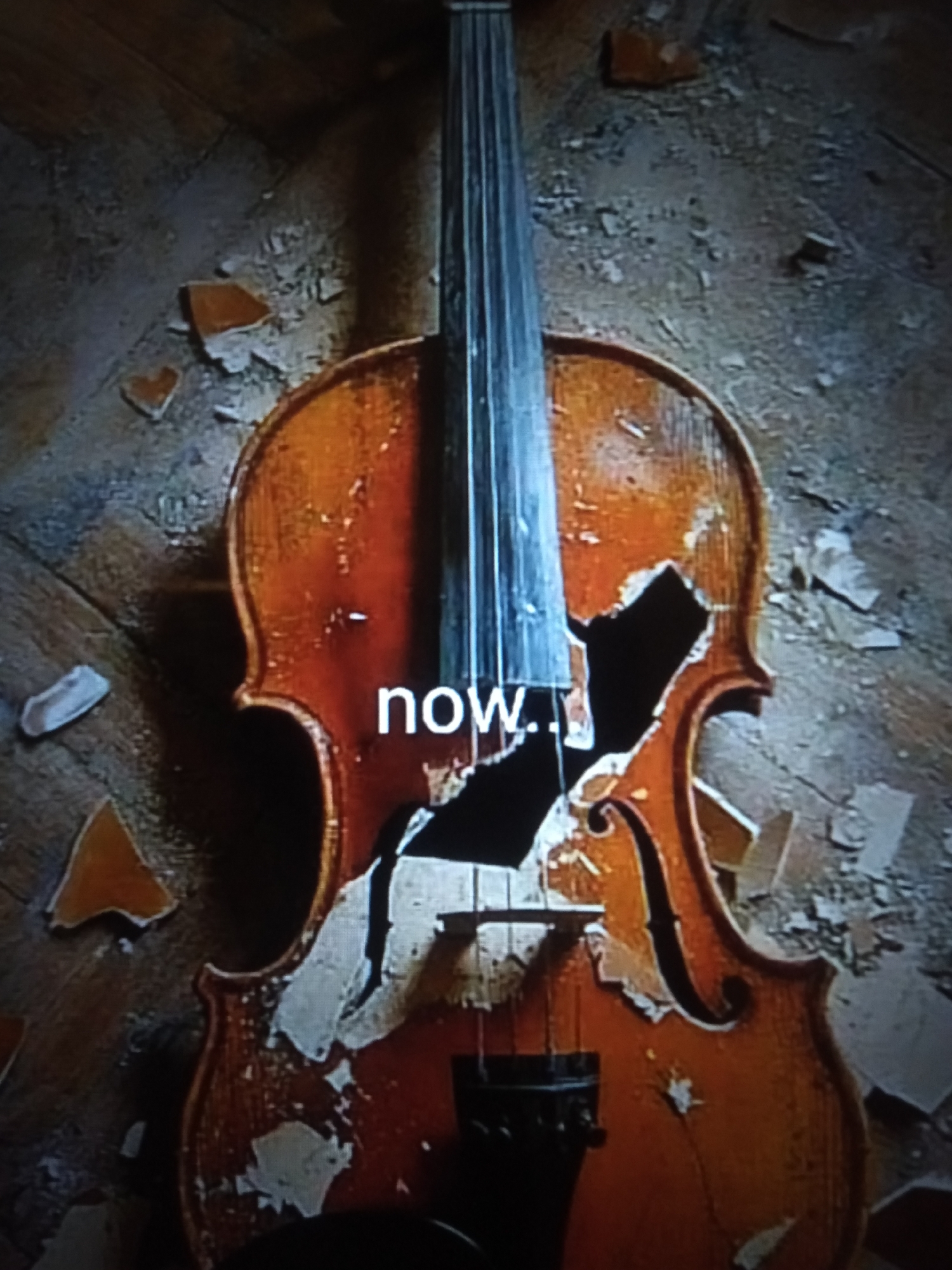 The image shows a broken cello lying on a wooden floor. The cello is cracked and pieces of wood are scattered around it. The word "now." is written in white text across the body of the cello. The scene appears to be indoors, possibly in a room with peeling wallpaper. There is no indication of the location in Jalingo, Nigeria.