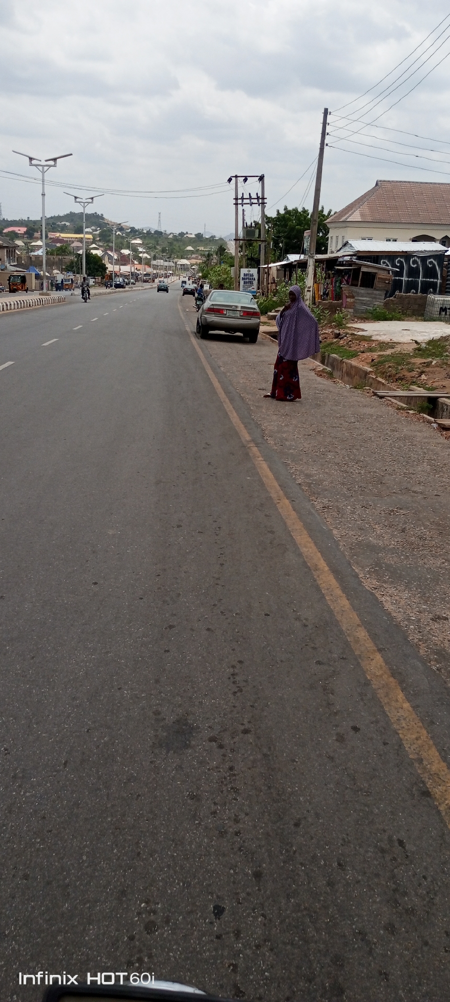 A street scene in Zango-ndaji, Nigeria is captured on an overcast day. A woman in a dark red dress and lavender head covering stands on the side of the road near a parked grey car. The road stretches into the distance with a yellow line marking the side. Other cars and motorcycles are seen in the distance. Buildings and houses line the street and a hill is visible in the background. Street lights and utility poles are visible along the road. The road surface is asphalt. The camera is an Infinix HOT 60i.
