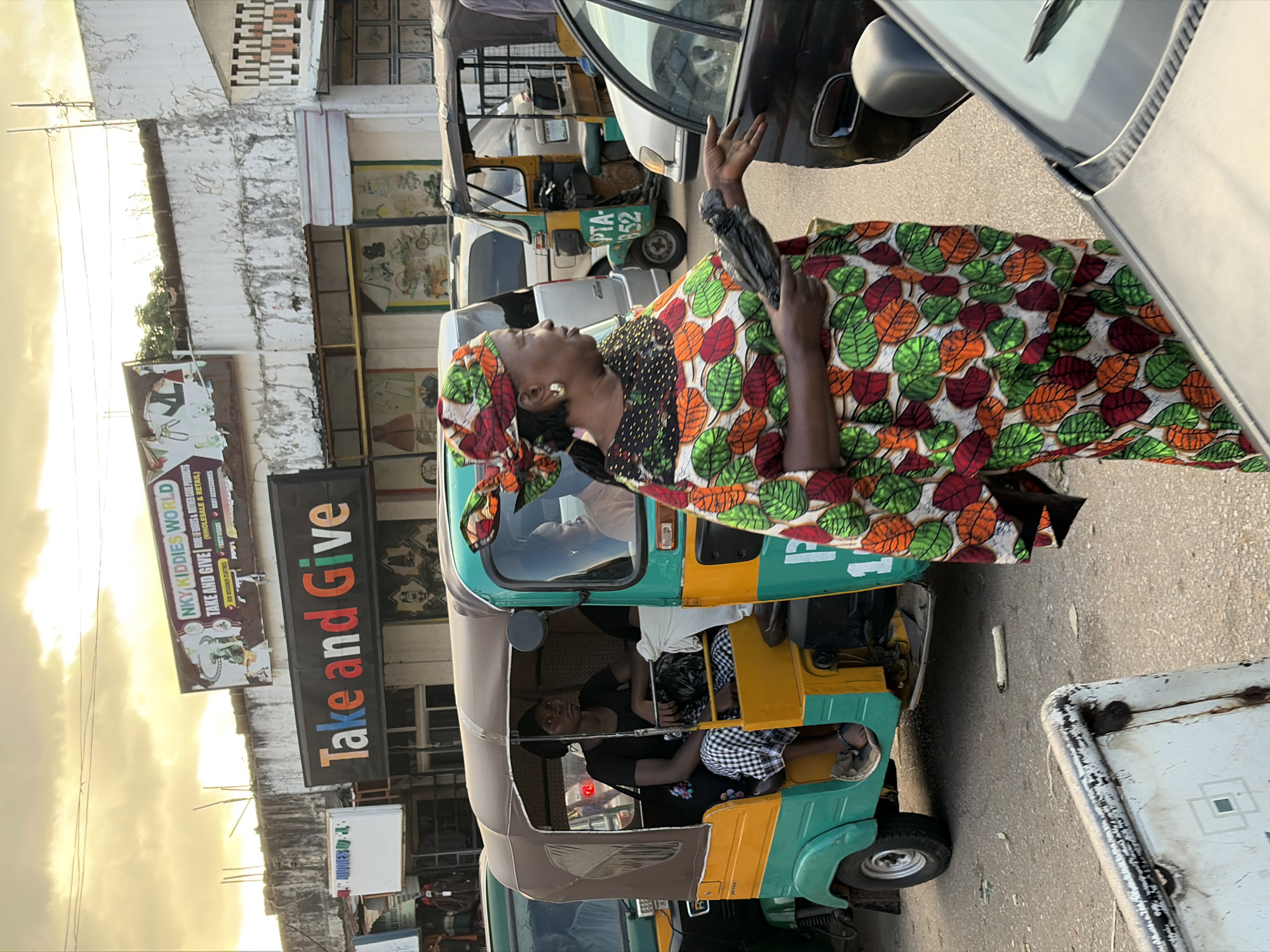 The image captures a street scene in Jos, Nigeria, during what appears to be late afternoon. A woman, dressed in a vibrant, leaf-patterned traditional outfit and headscarf, stands on the right side of the frame, facing a dark-colored car. She holds a dark plastic bag in her left hand and extends her right hand, palm open, towards the car, suggesting an interaction or transaction.
In the background, a green and yellow auto-rickshaw, a common mode of transport in Nigeria, is parked or moving slowly. A woman wearing a black headscarf and a checkered skirt is seated in the passenger section of the auto-rickshaw, looking forward. Another person, partially visible, is in the driver's seat. Further behind, other vehicles and buildings line the street.
Above the buildings, a cloudy sky with warm tones indicates the setting sun. A large sign on one of the buildings reads "Take and Give" in large, colorful letters, and another smaller sign above it says "NKY KIDDIES WORLD." The environment appears to be a commercial area with shops and pedestrian activity. The overall atmosphere suggests a busy urban setting.